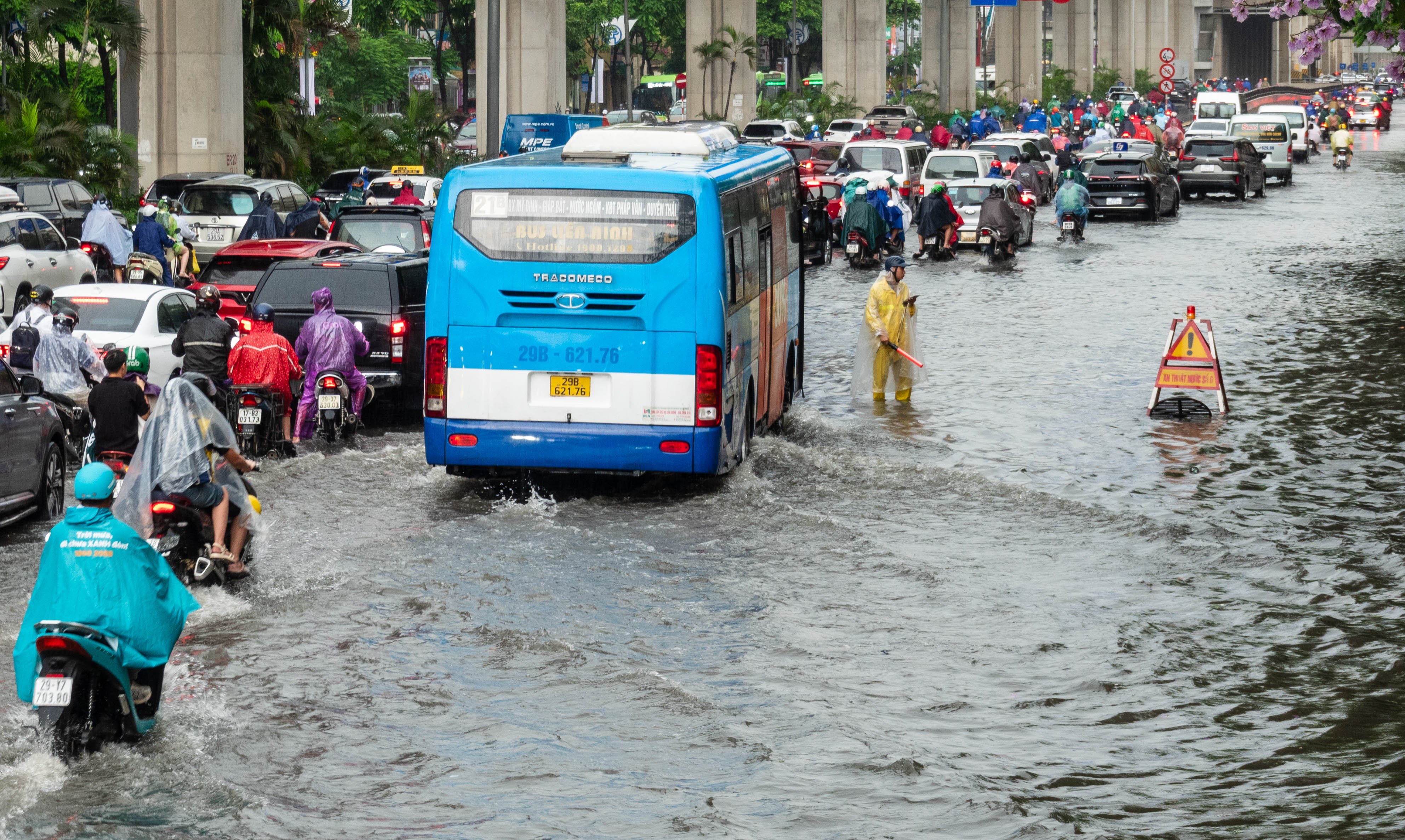 Traffic in deep water. Blue bus in foreground. Man in yellow standing in the water near a warning sign.