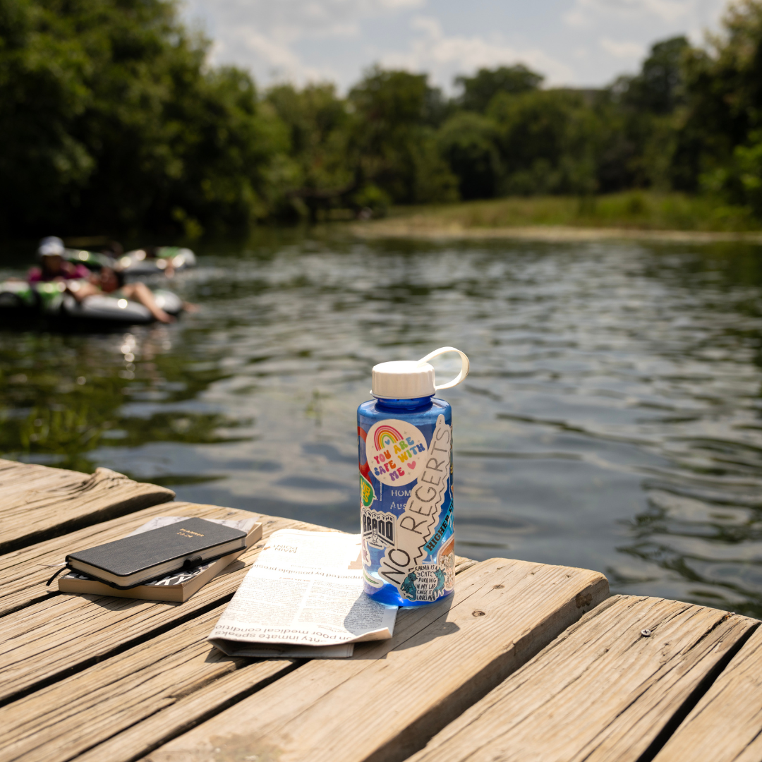 Water bottle covered in stickers with a notebook and newspaper nearby. Located on the dock at Sewell Park with a scenic view of the San Marcos River in the background with two river tubes float by in the distance.