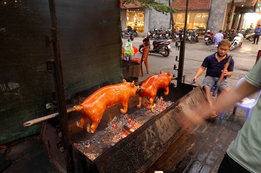 two piglets roasting over an open pit. the cook is on the right hand turning the roasting crank with one hand while holding a 28" wooden water pipe for smoking tobbaco. 