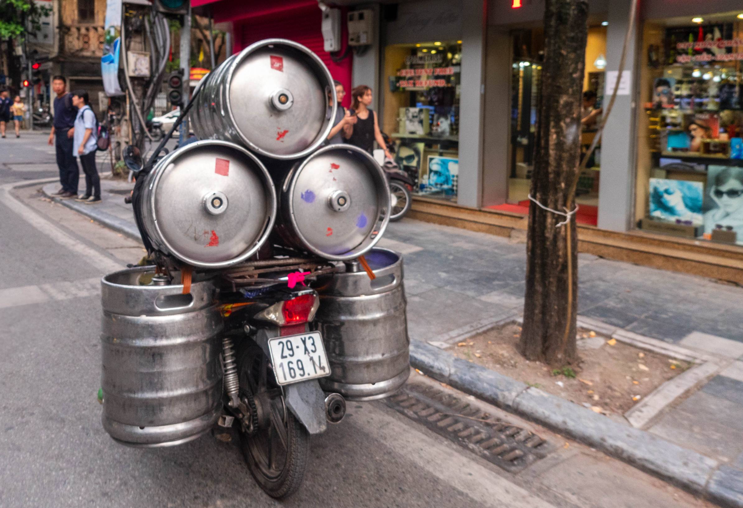 Five 16-gal steel kegs strapped on a motor bike driving down road. 