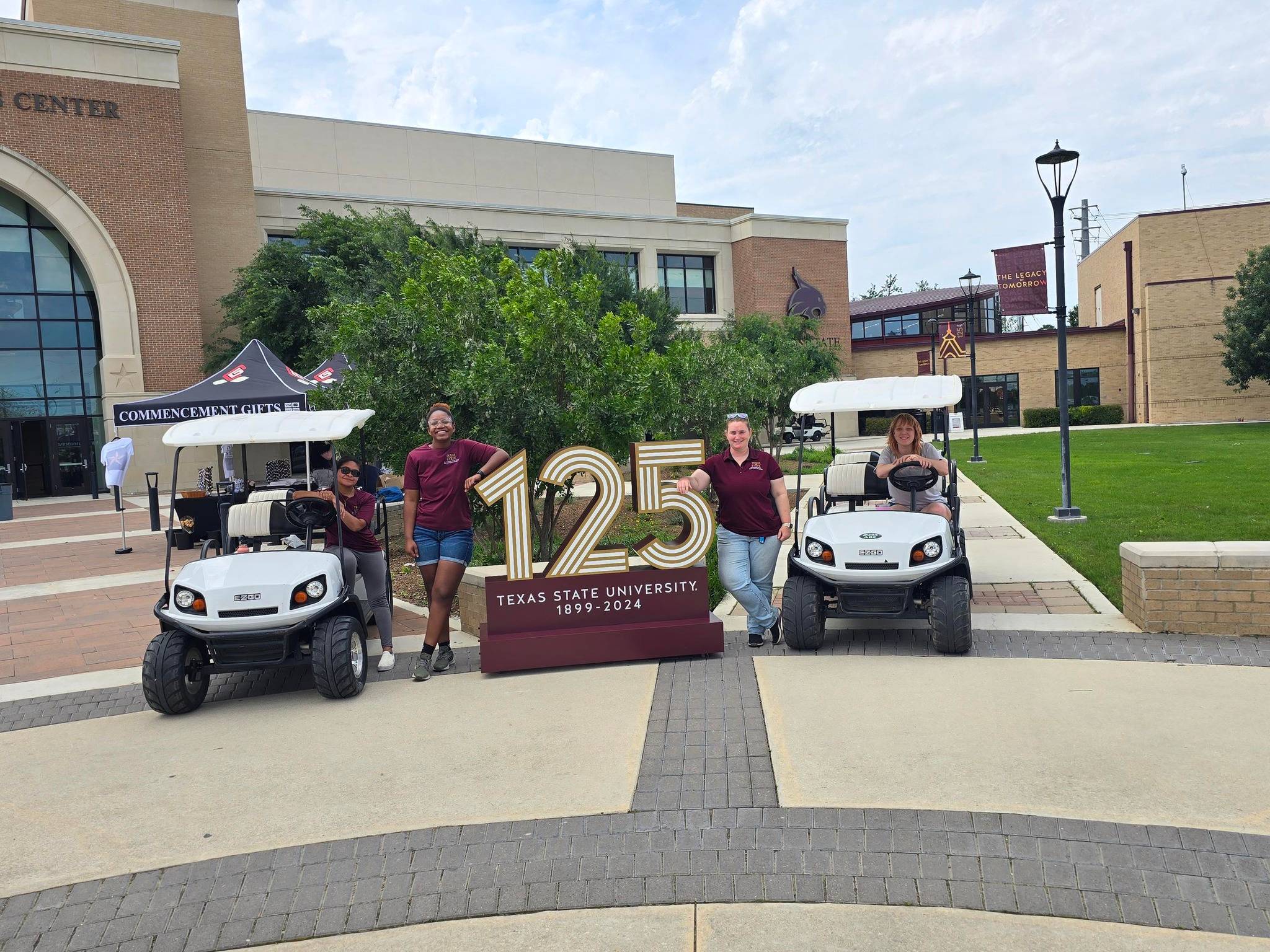 Two golf carts parked with 125 year sign in middle with one student on each golf cart and one student of the left side of the 125 year sign and a staff member on the right side of the 125 year sign