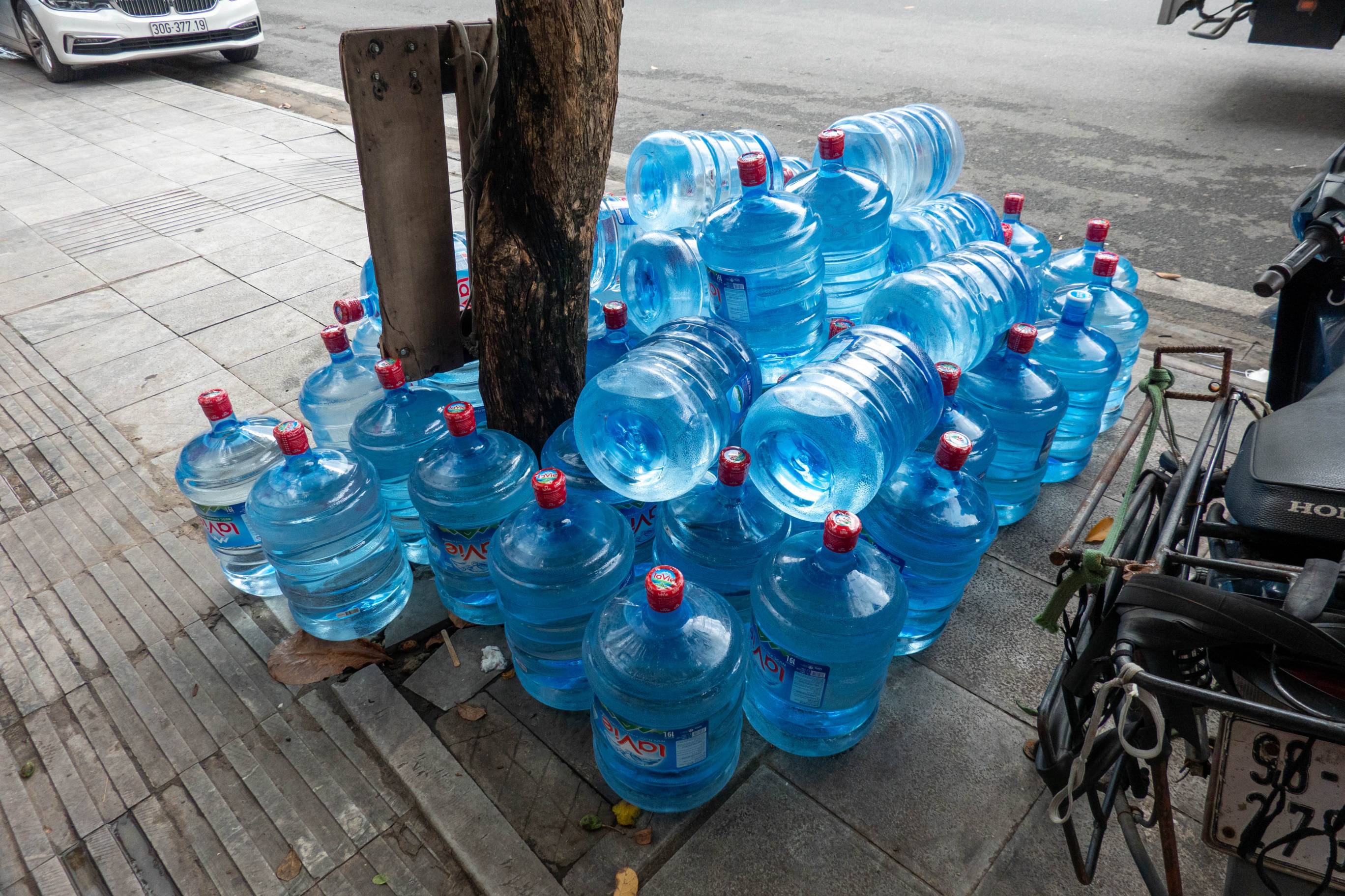 Unopened blue 5 gallon water jugs piled up on sidewalk around a tree trunk next to a street.