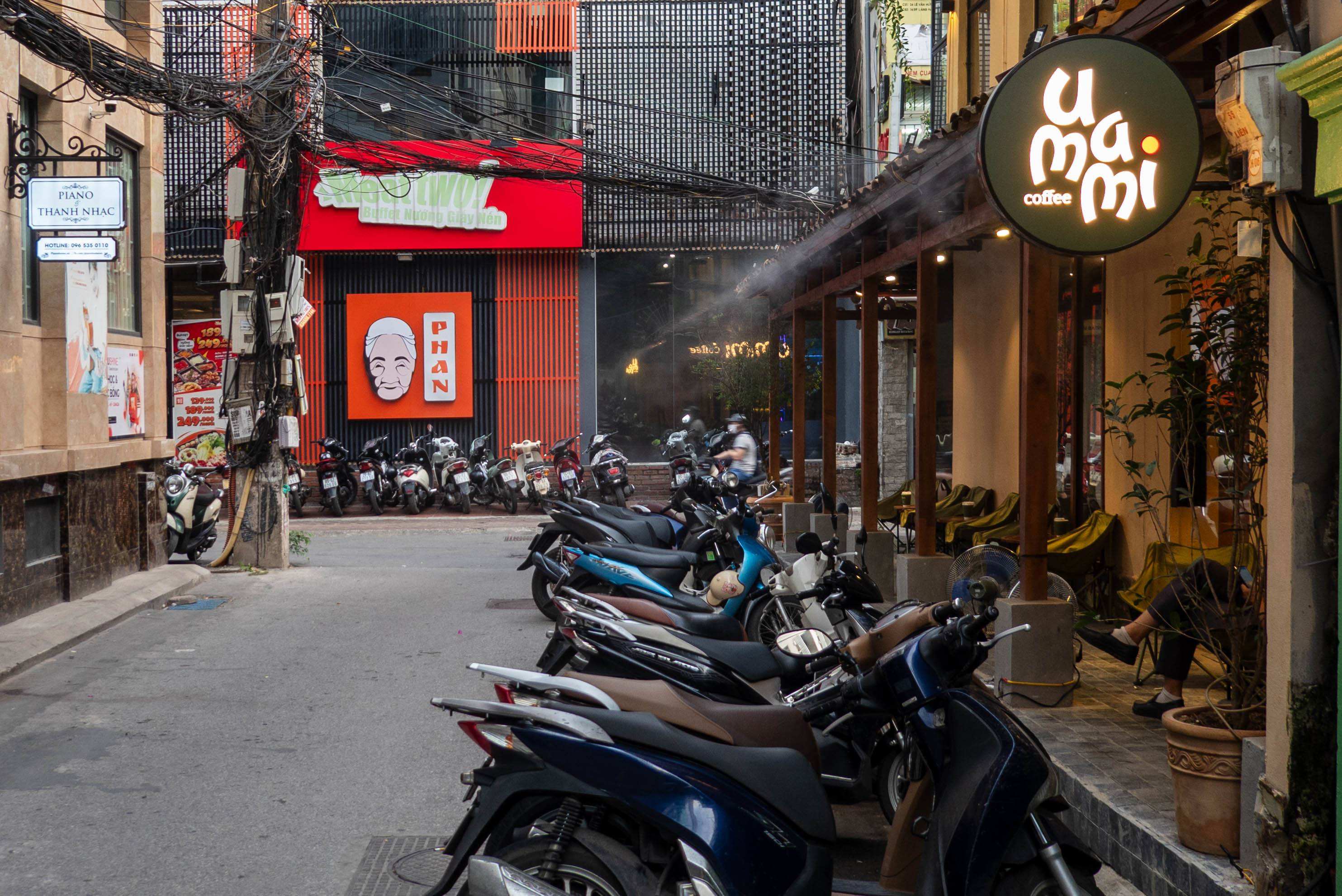 building on right is the Uammi Coffee house. Up by the rain gutter is spraying water mist. There are motor bikes in front of the coffes shop. at the end of the alley is another business called Phan (orange sign) with motorbikes in front of it.