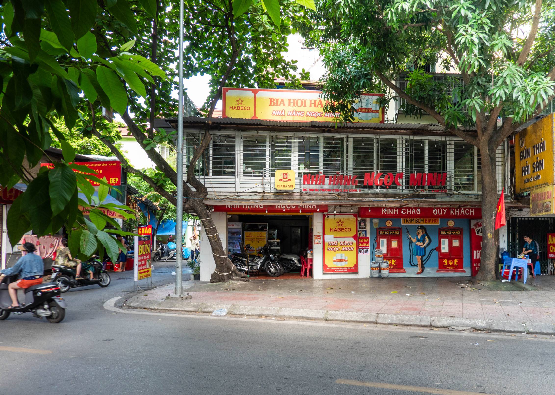 picture of building on street corner. Glass inclosed area  on top and a picture painted on wall in lower right showing a woman standing betwen two gas pumps that say bia hoi.