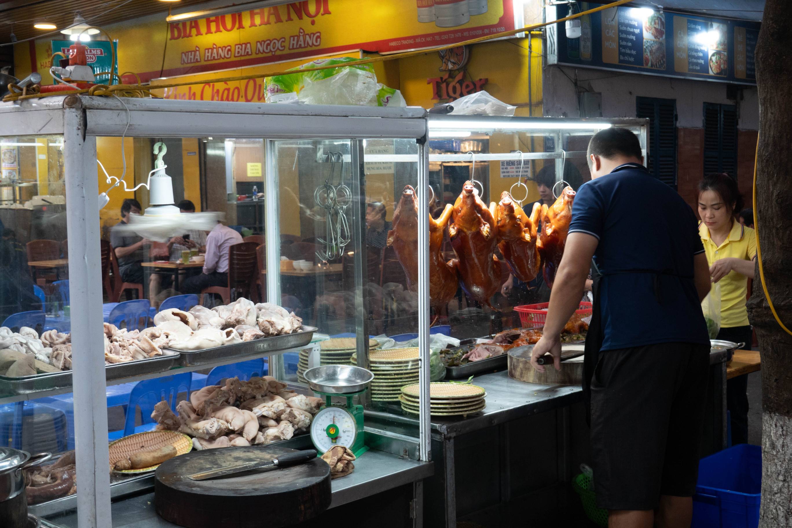 picture of food kiosk. On left are pieces of boiled meat. On the right a man is standing in front of four roasted pigs hanging from a bar. He is choping up food. A woman in a yellow shirt stands beside him to his right.  Part of the top of a storefront is visiable above the kiosk.