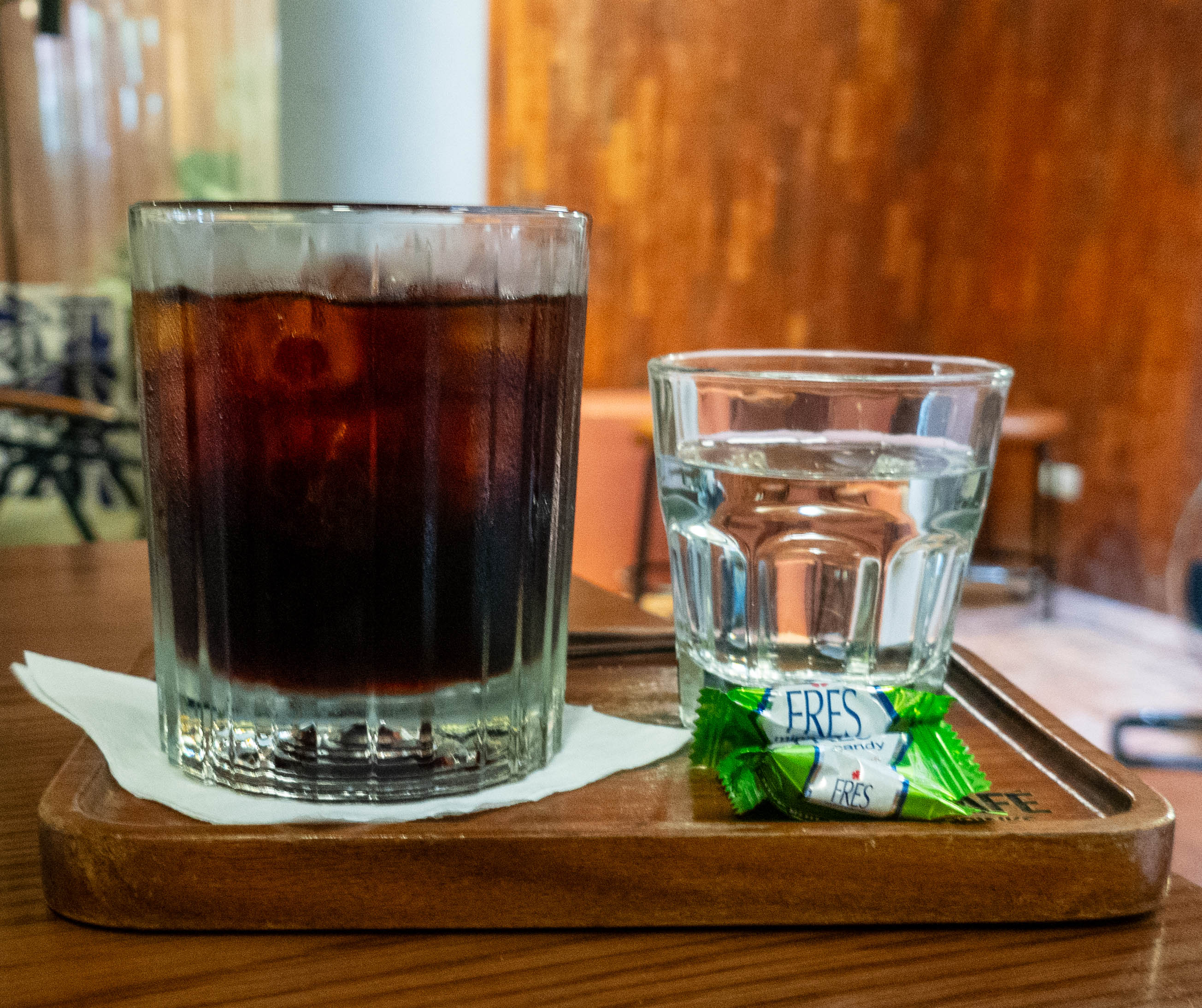 glass of ice coffee with small glass of water to the right. The coffee now has a small white paper napkin under it.