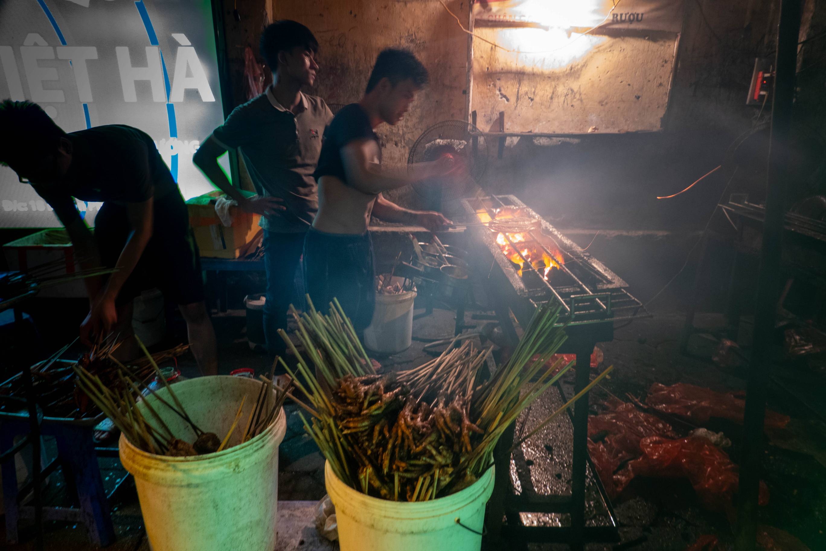 man cooking pieces of chicken on open pit in background. In foreground there is a five gallon bucket full of roasted chicken feet on wooden skewers.