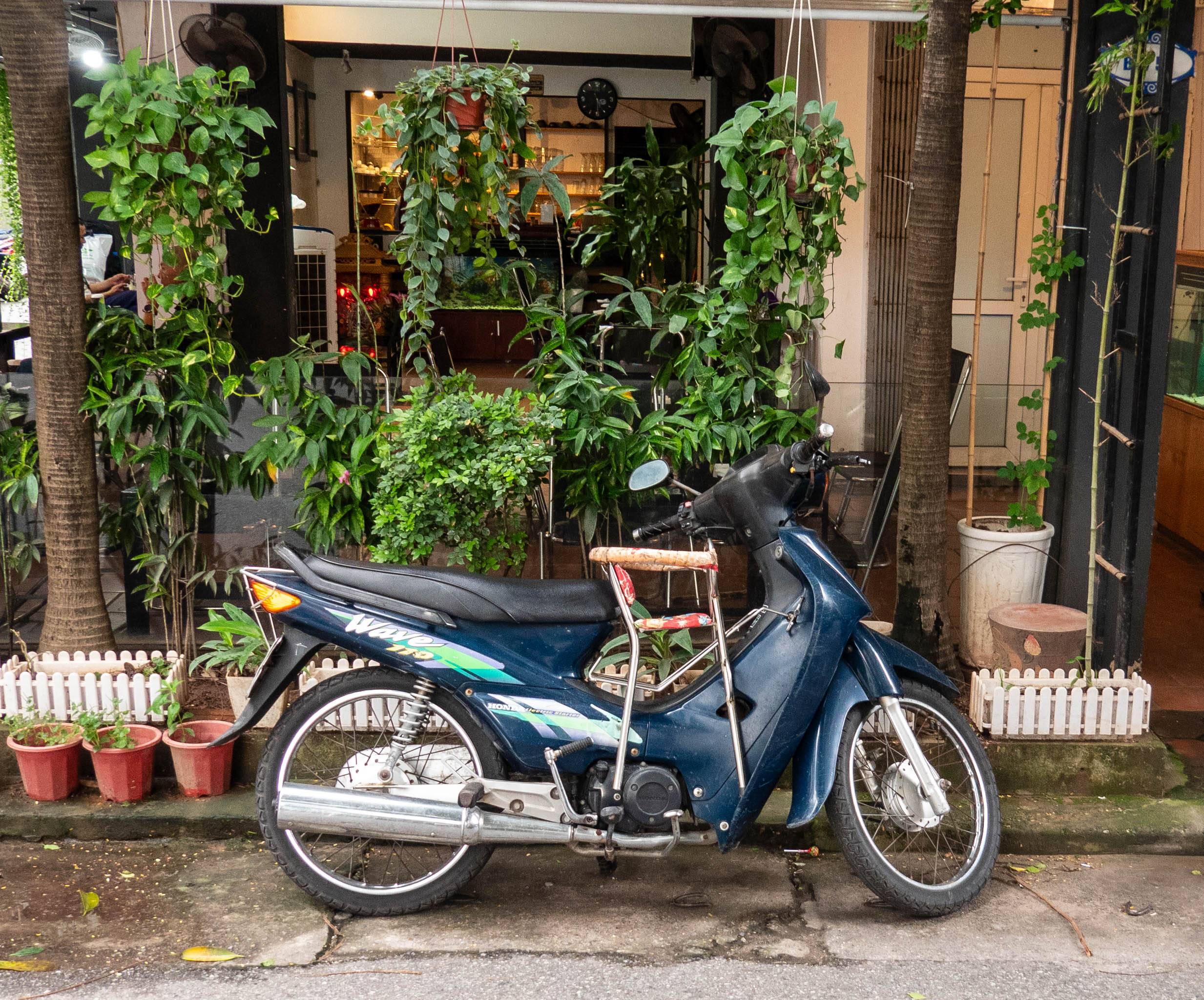 picture of motorbike in front of coffee shop.  There is a metal highchair strapped to the motorbike between the driver and the handlebars of the motorbike