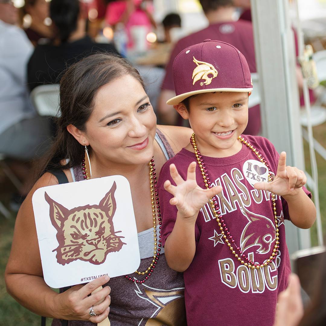 A mom and her little kid smiling for a photo wearing Texas State merch at an alumni tailgate