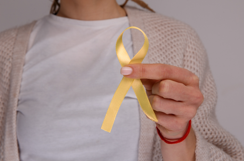 a person in a white shirt and beige cardigan holding a gold awareness ribbon