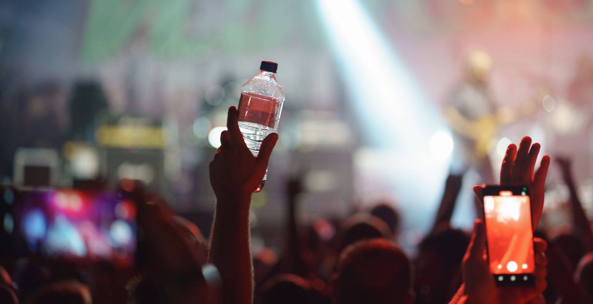 An image of someone's hand holding a water bottle up in the air with a concert performance happening in the background.