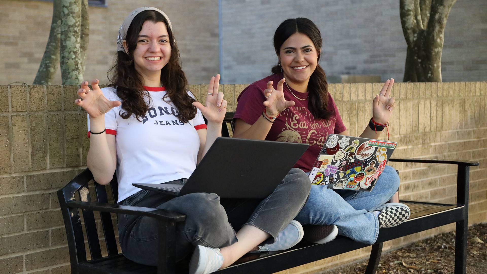 Two students on a bench studying on laptops, talking, and smiling