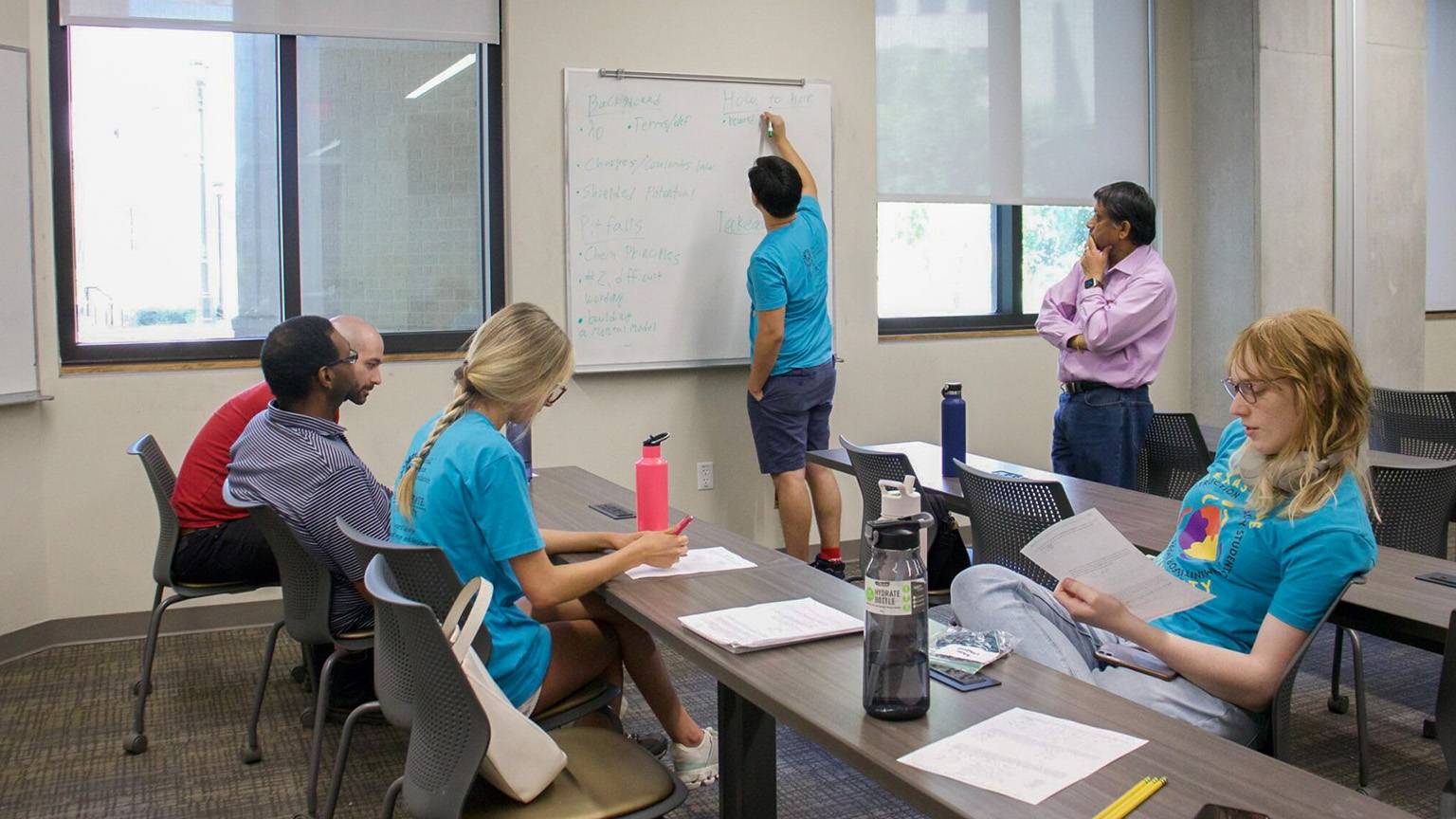 A student writes something on a white board while other students take notes.