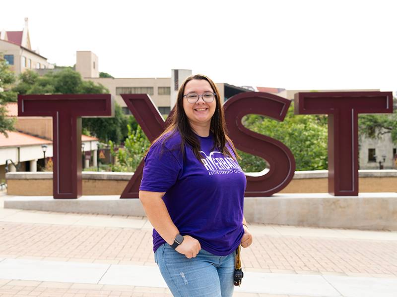 female student in ACC Riverbats t-shirt posing in front of TXST letters sculpture on campus