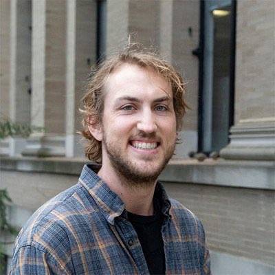 man with beard wearing plaid shirt and dark undershirt in front of building