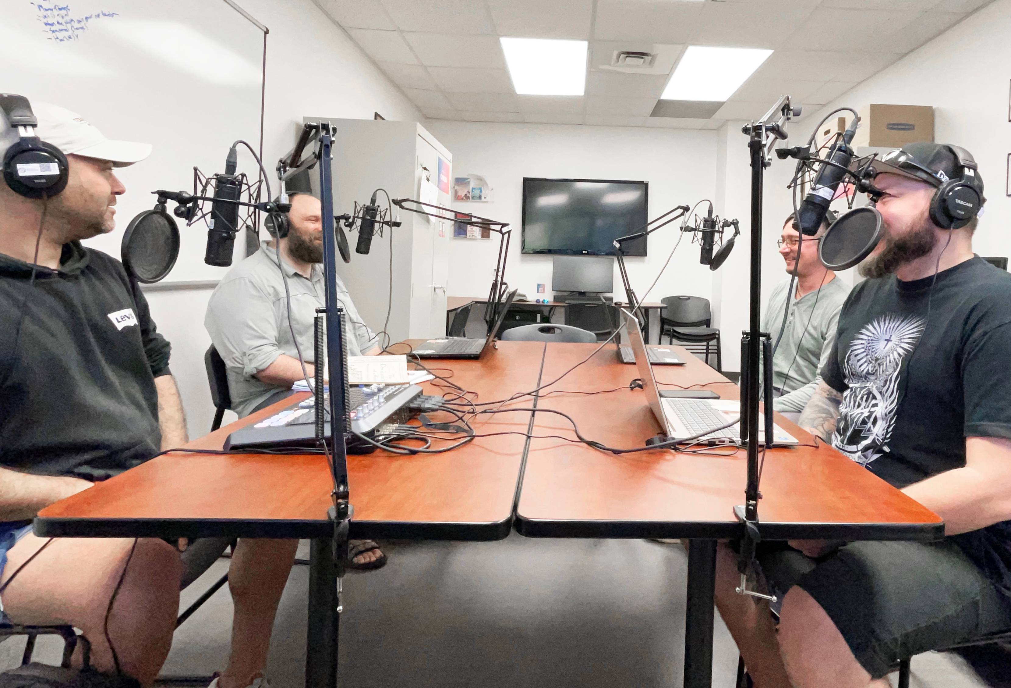 Four men sit talking around a table covered in podcasting equipment and laptops.