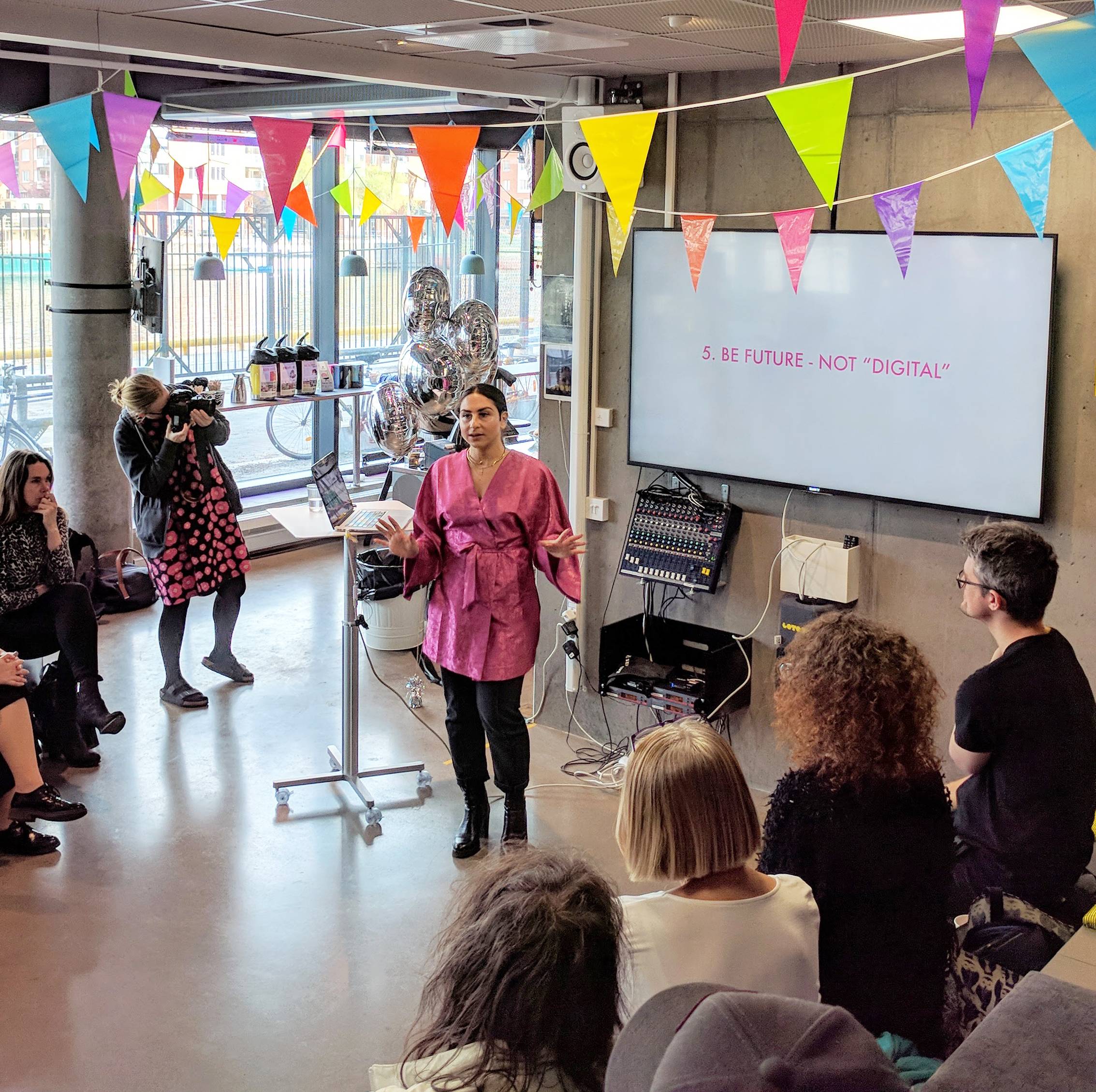 A woman presents to a crowd in a festively decorated space. A screen behind her says "Be future Not Digital"