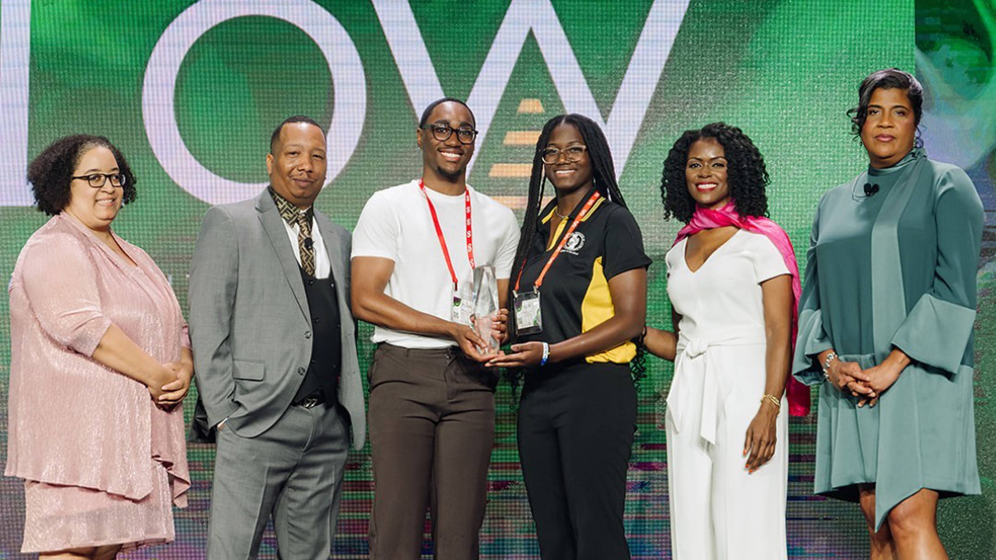 Two college students accepting award at center, flanked by two event officials on each side