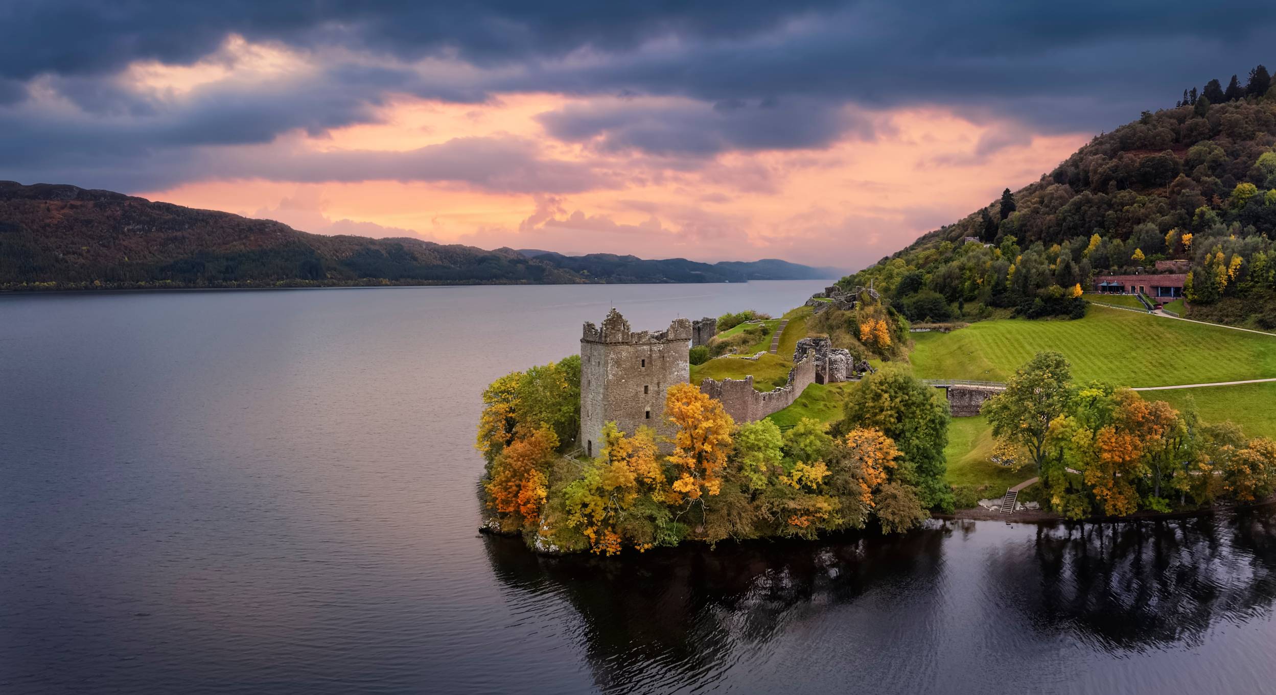 Historic Urquhart Castle perched on the shores of Loch Ness, illuminated by the warm hues of sunset, with the tranquil waters reflecting the colorful sky."
