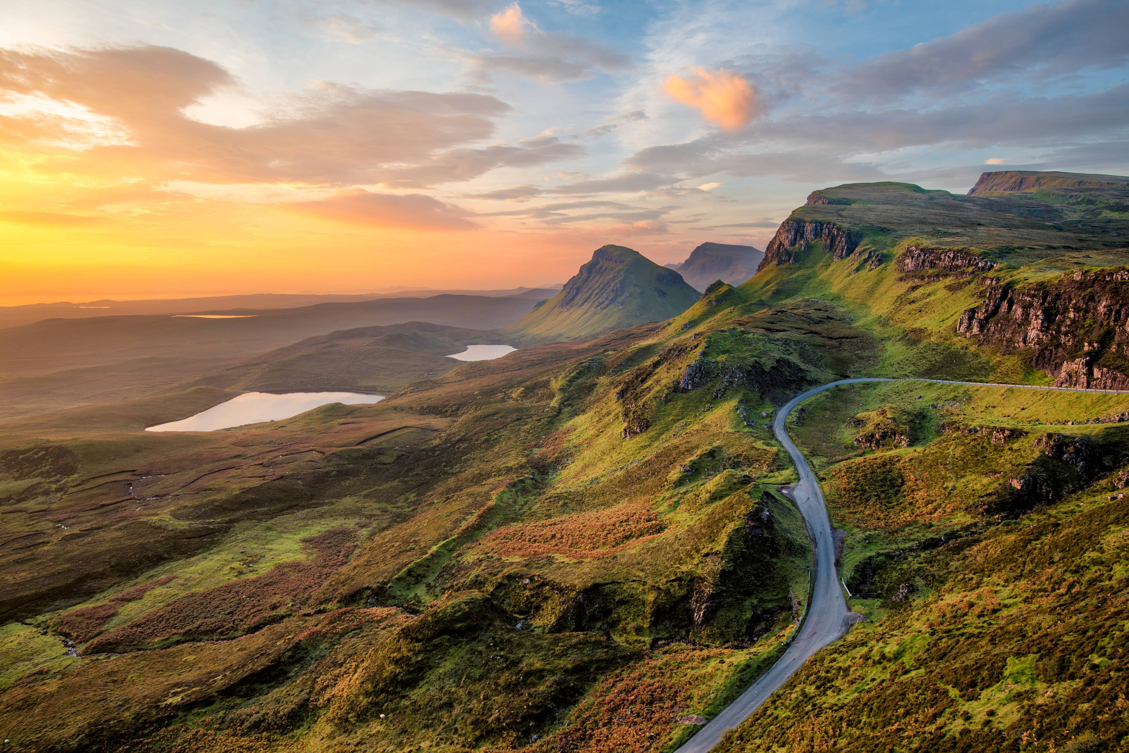 A golden sunrise casting over the Isle of Skye and the rugged Cuillin Hills, while a winding road cuts through the landscape, inviting exploration.