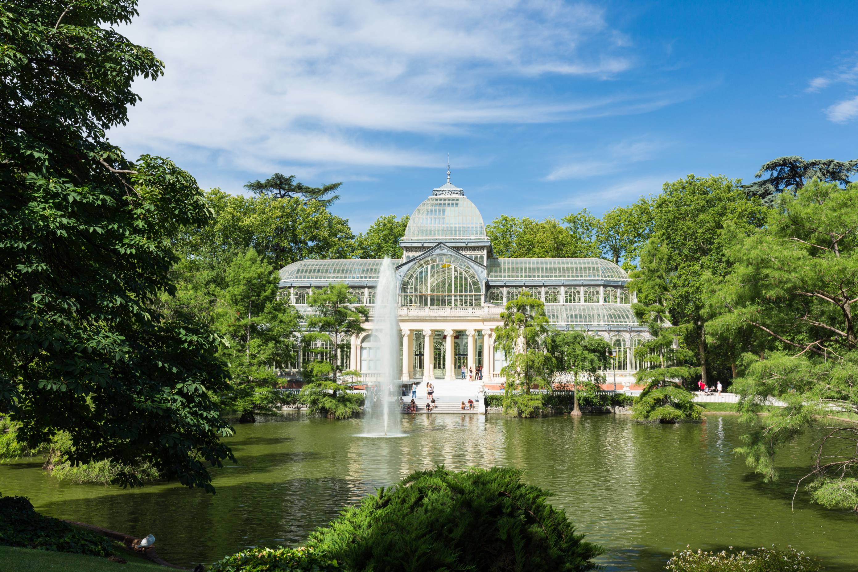 The Glass Palace, viewed from a distance in Retiro Park, is framed by lush greenery, with a tranquil pond and an elegant fountain in the foreground.