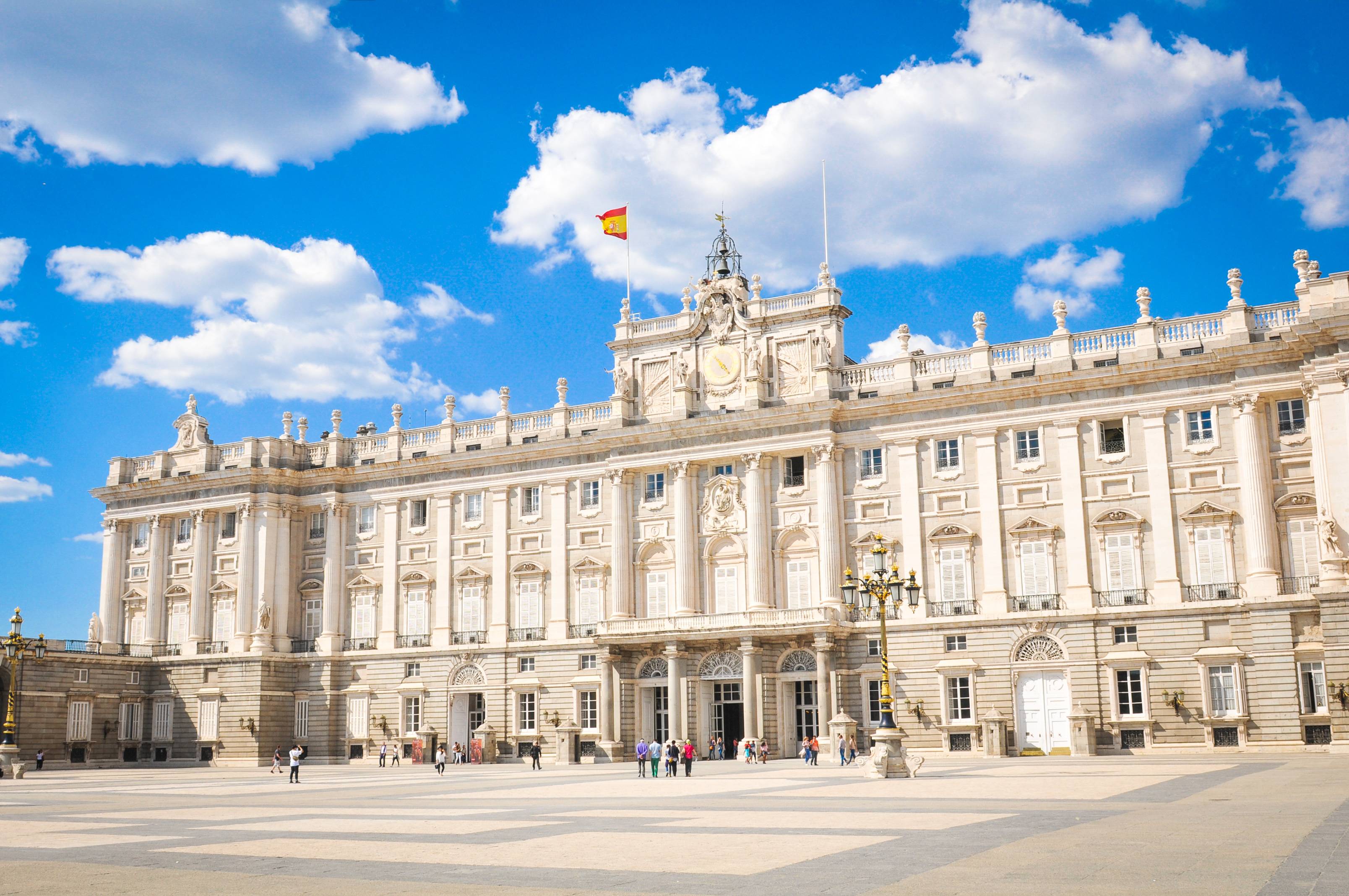 Outside view of ornate white building with flag of Spain and bells with pedestrian onlookers.