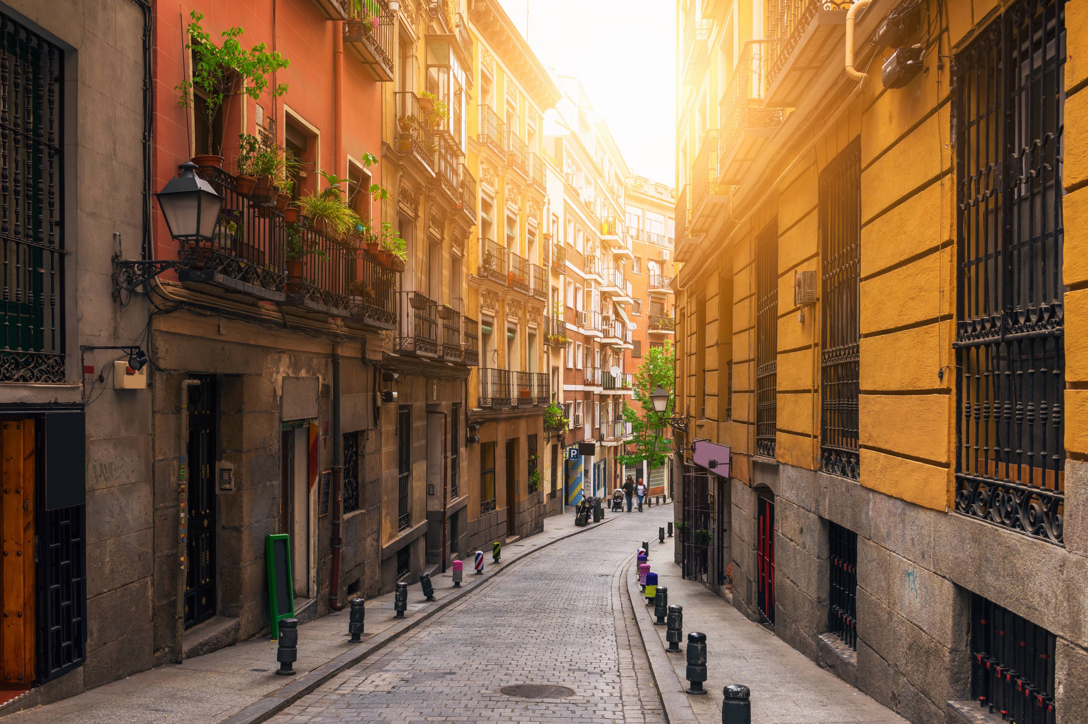 view of a narrow stone street in Madrid with sunlight filtering through gap between tall connected residential buildings.
