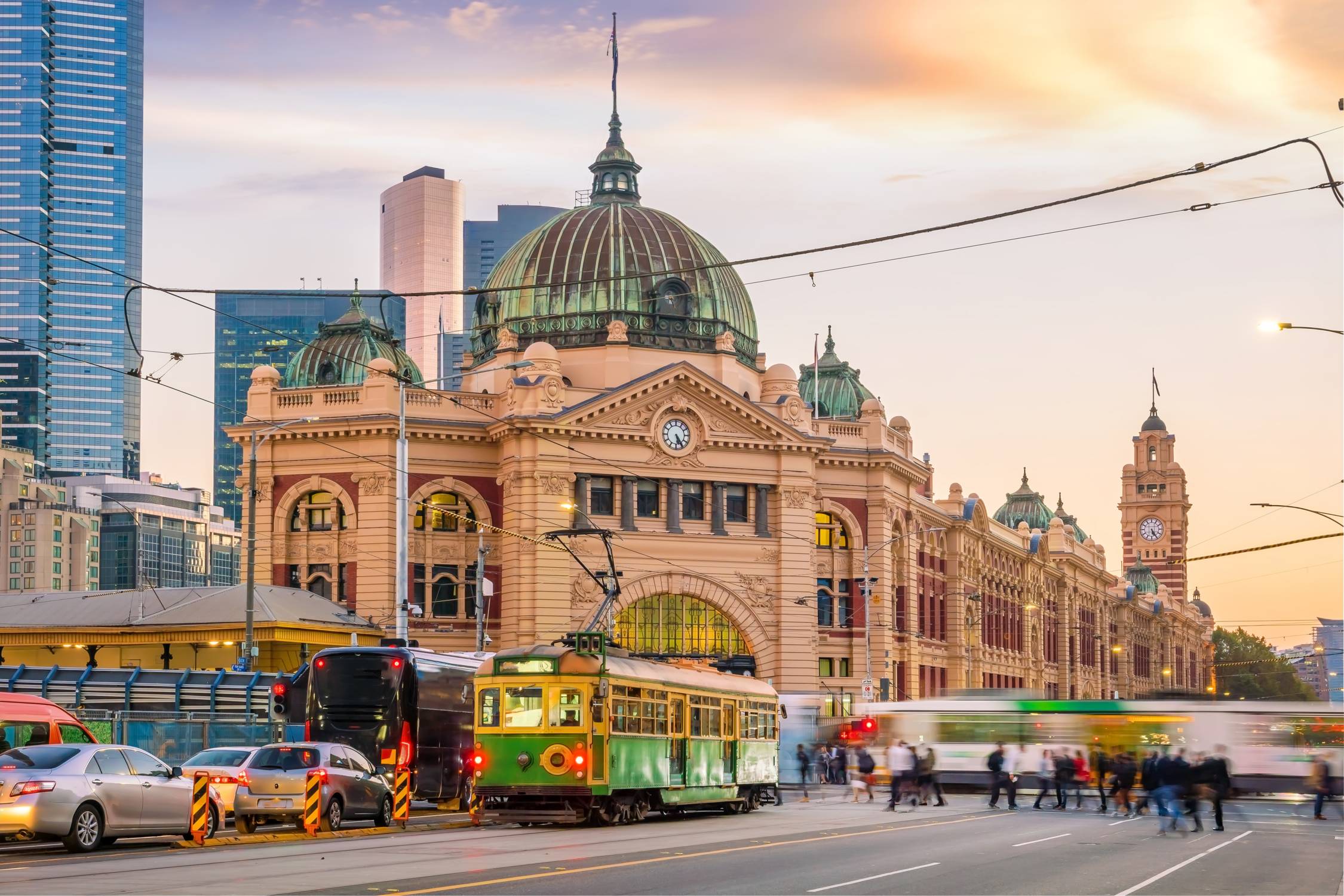 View of historic Flinders Street Railway Station, with trollys and traffic in front, set against a backdrop of modern architecture.