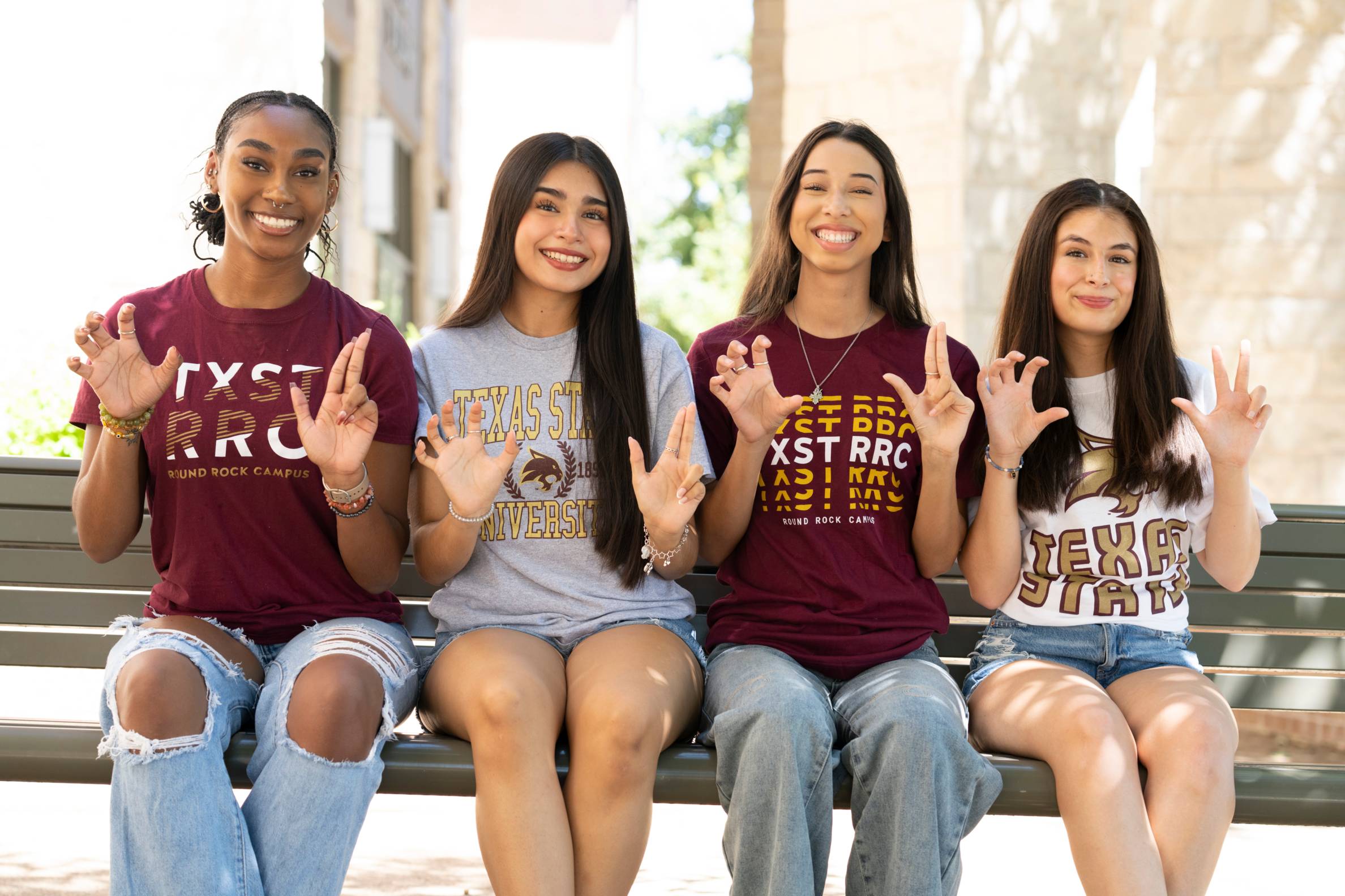 four young women sit on a bench holding up TXST hand signs