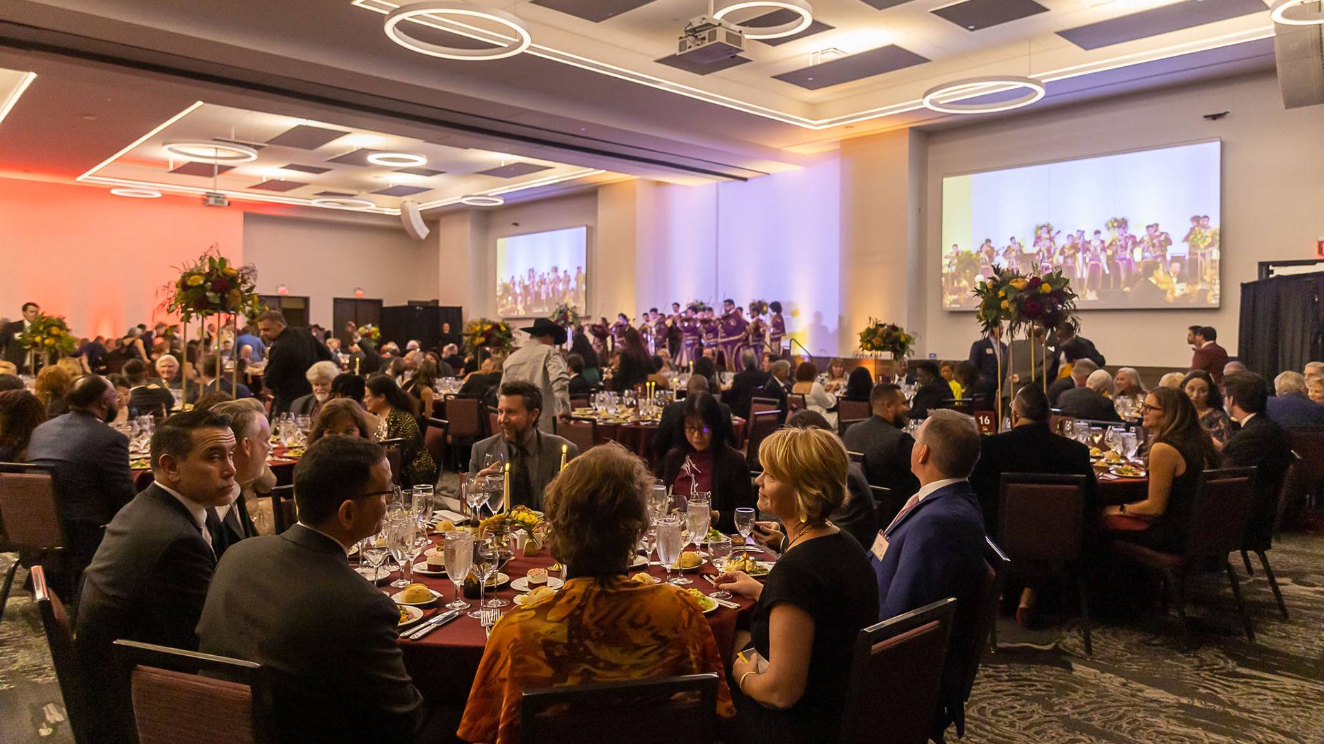 Tables of smartly dressed people seated in a large room, discussing over elaborately laid out food and drink.