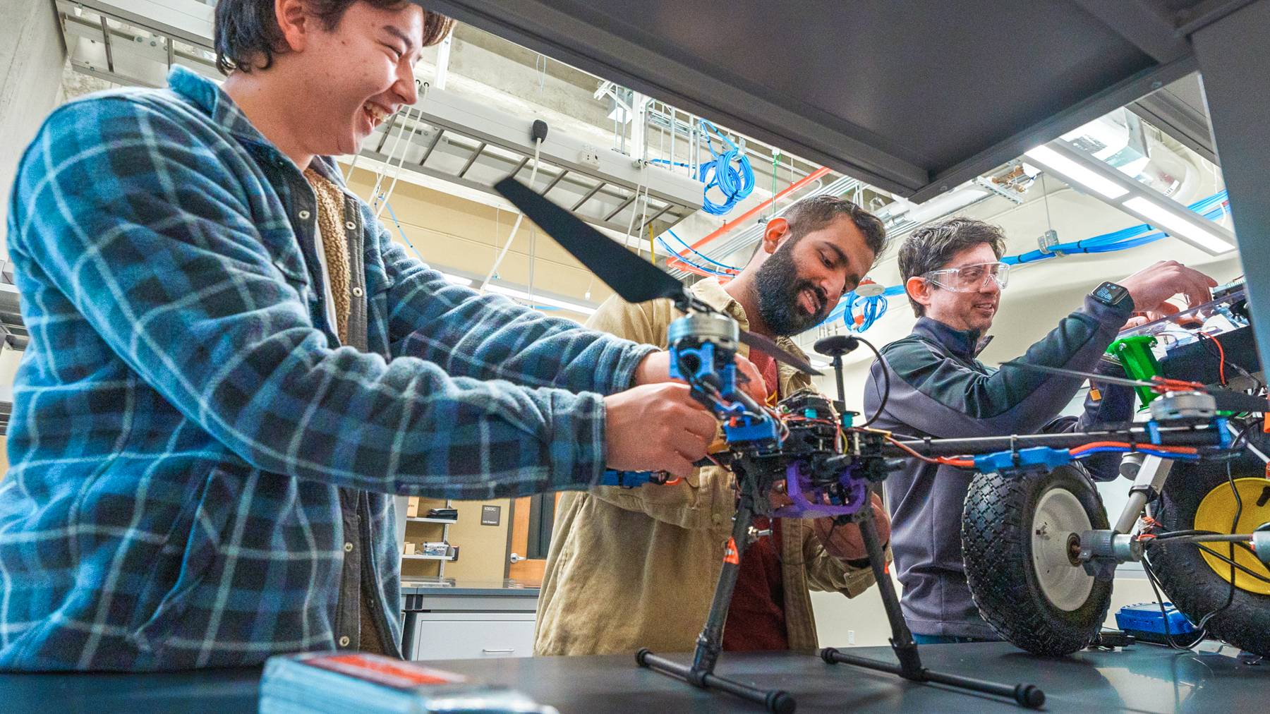 A professor and two students work on two engineered drones and a rover at a lab.