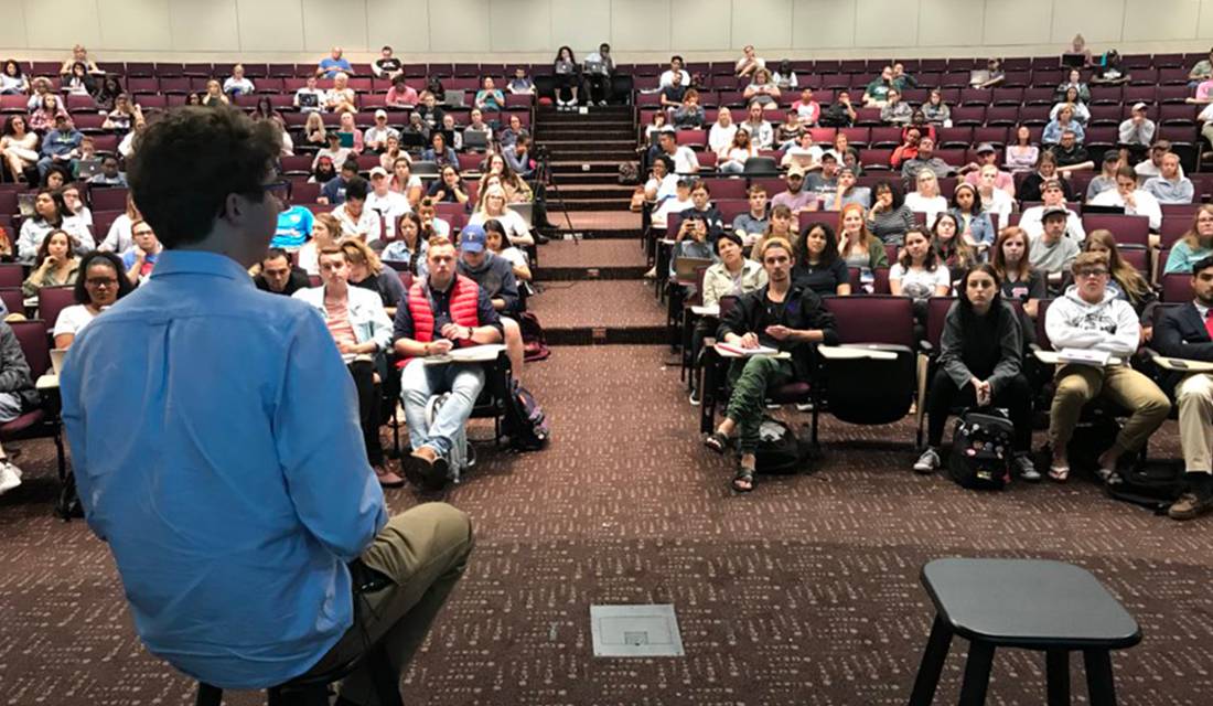 Man in a blue button-up shirt speaking in front of a classroom of students.