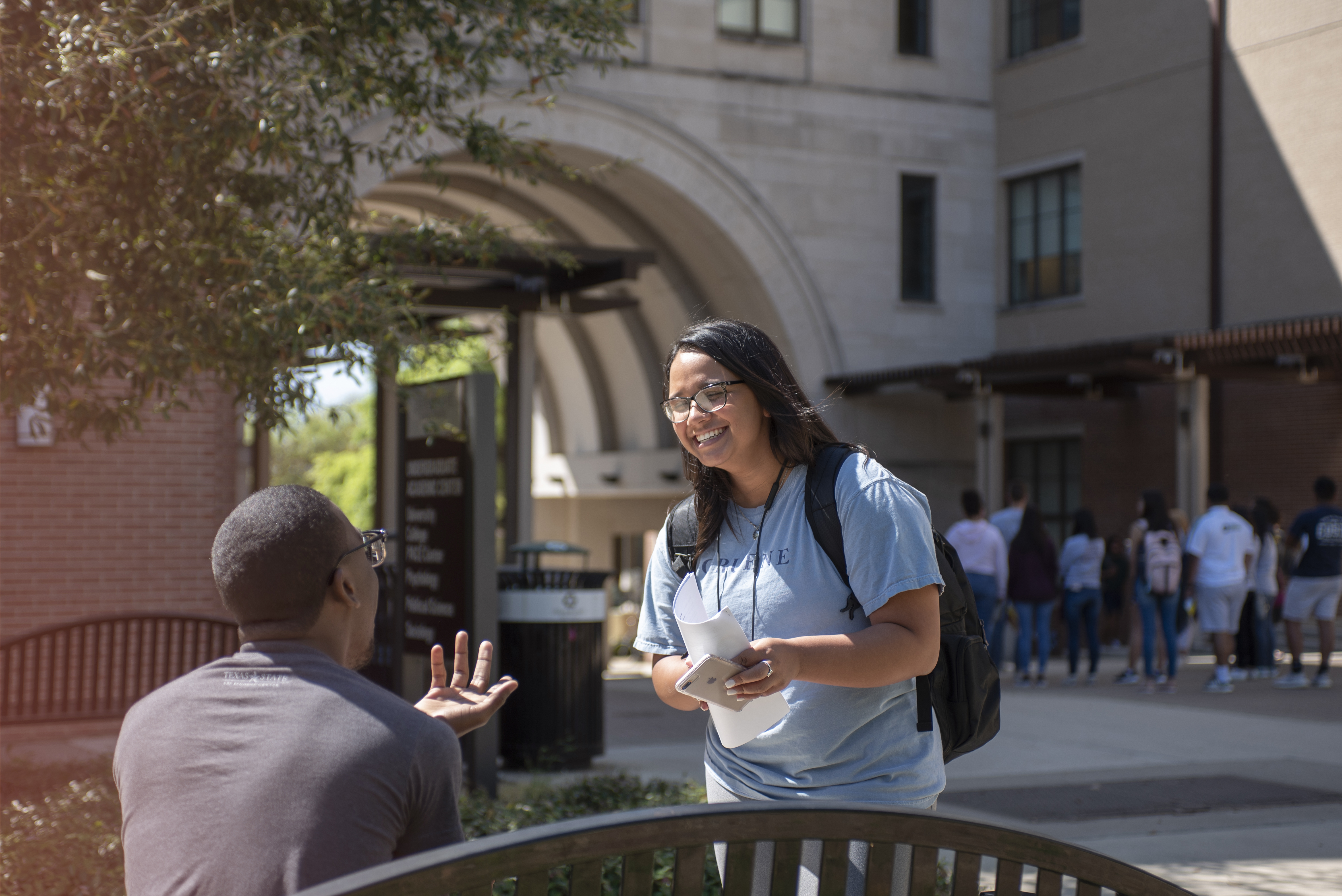 two students conversing at campus bench with the Trauth Huffman arch and fellow students in the background.