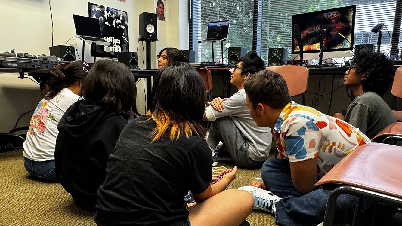 Students sit on the ground while listening to music through a sound system.