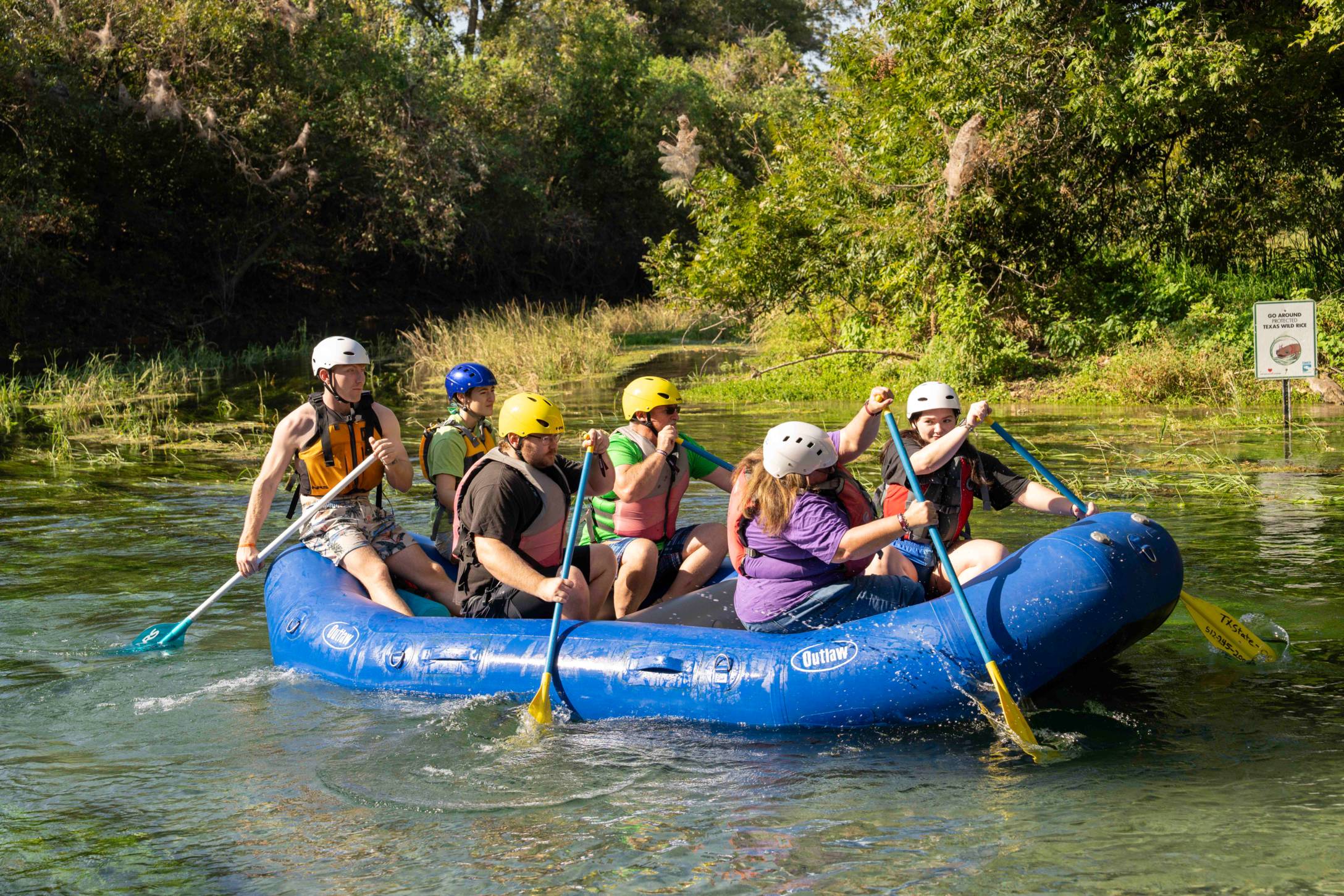 A group of people wearing life vests and helmets paddle in a raft on a river.