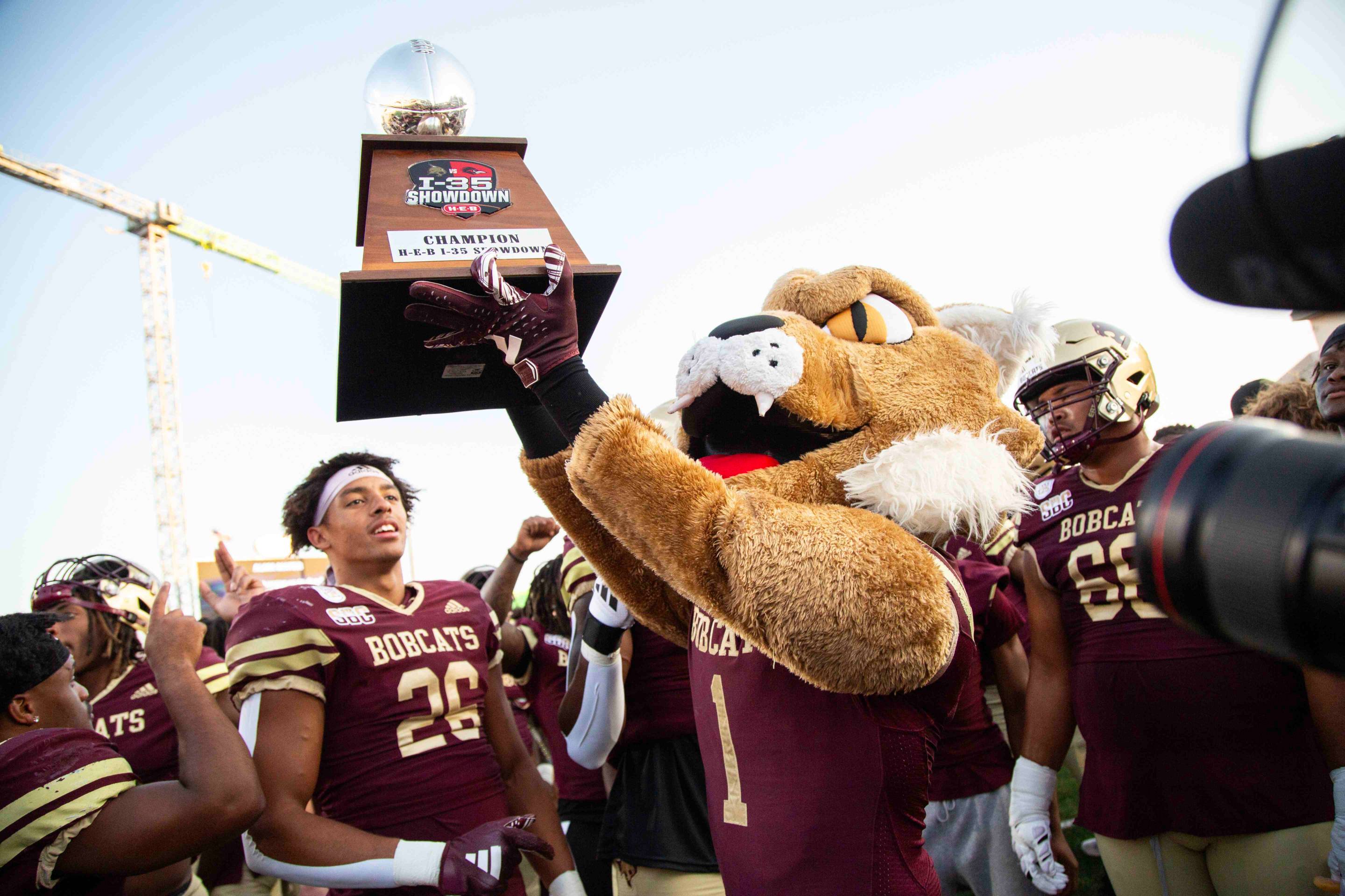 Boko the Bobcat holds a trophy aloft while football players in maroon uniforms celebrate below.