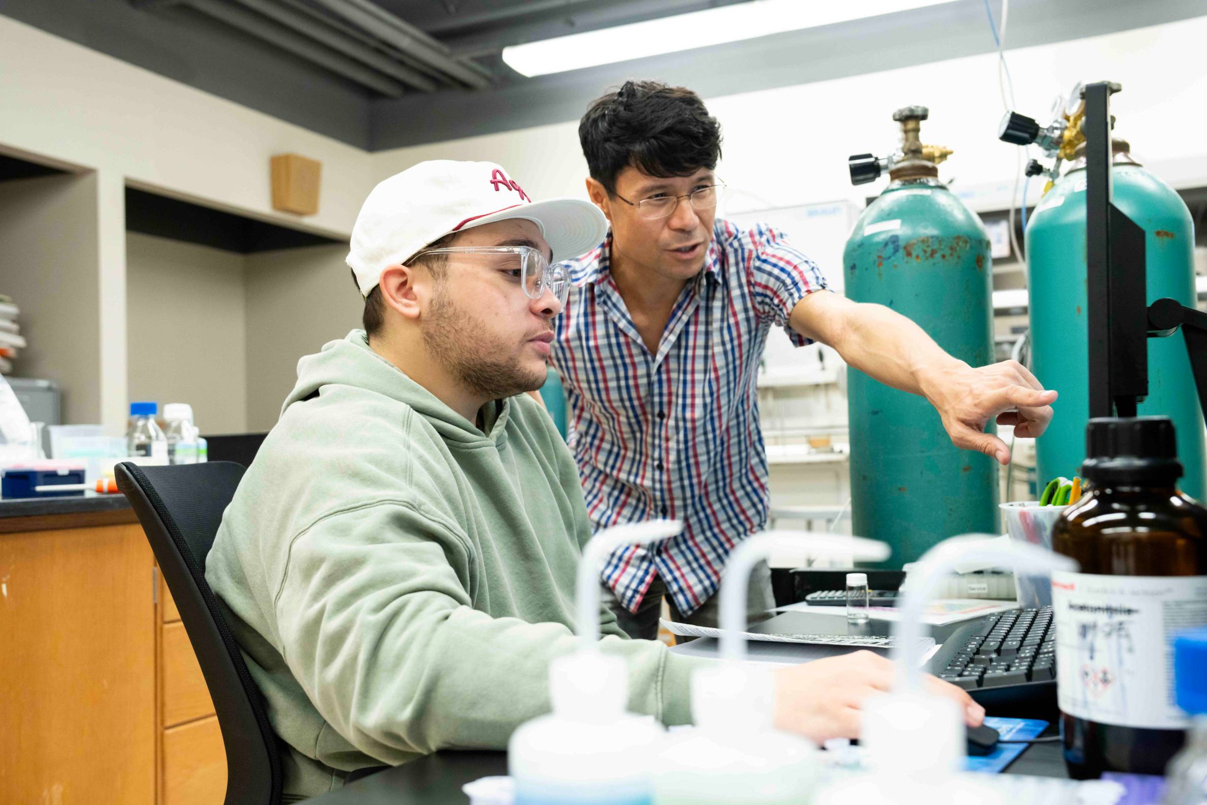 A professor in a laboratory setting points to something on a a student's laptop screen while the student looks on.