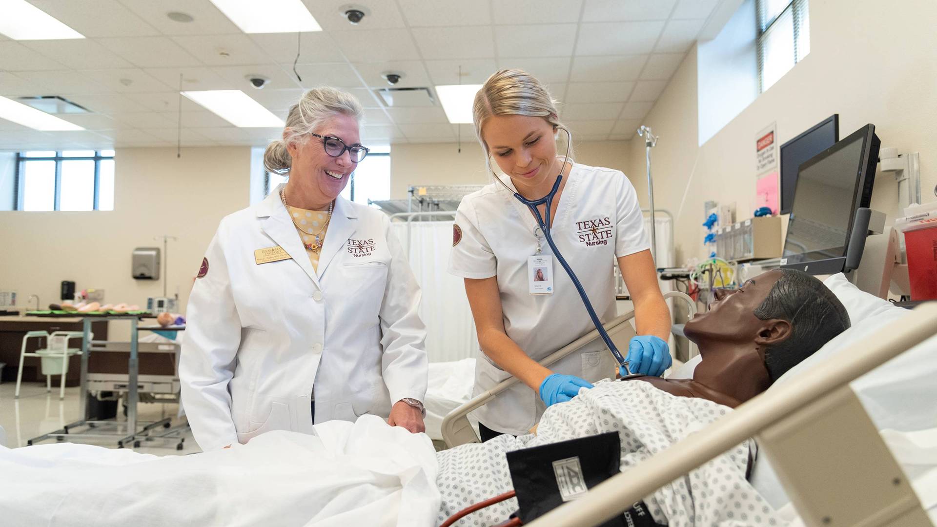 A nursing student practicing medical care in a nursing classroom with a professor wearing a Texas State nursing lab coat