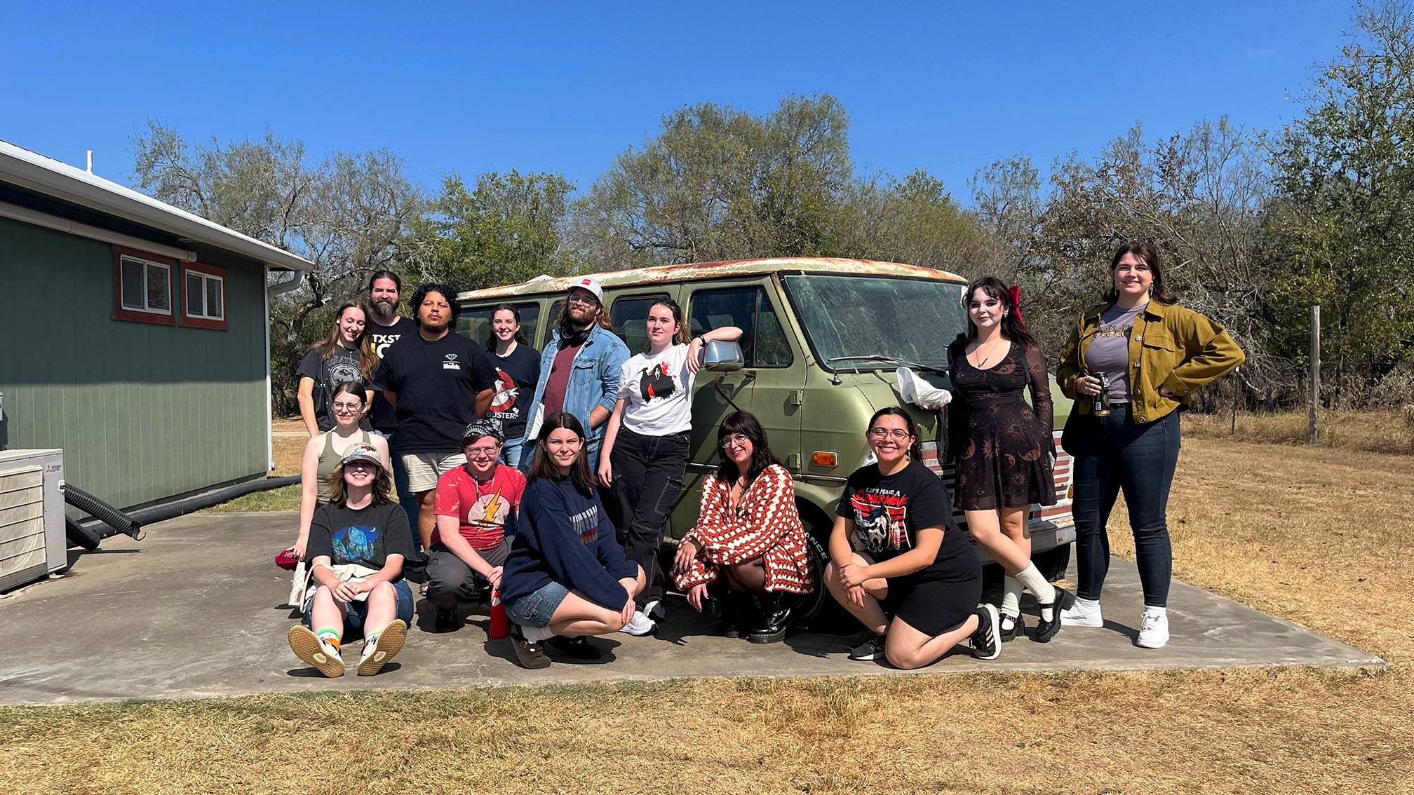 A group of students pose for a photo in front of a worn-down van at The Gas Station.