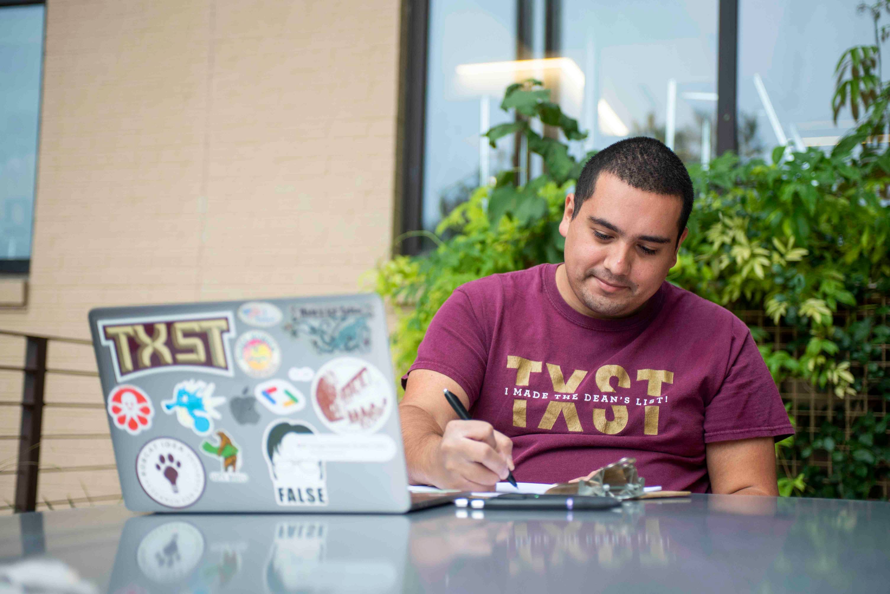 A young man wearing a maroon TXST shirt writes something down on a notepad. In front of him is a laptop, heavily covered in various stickers.