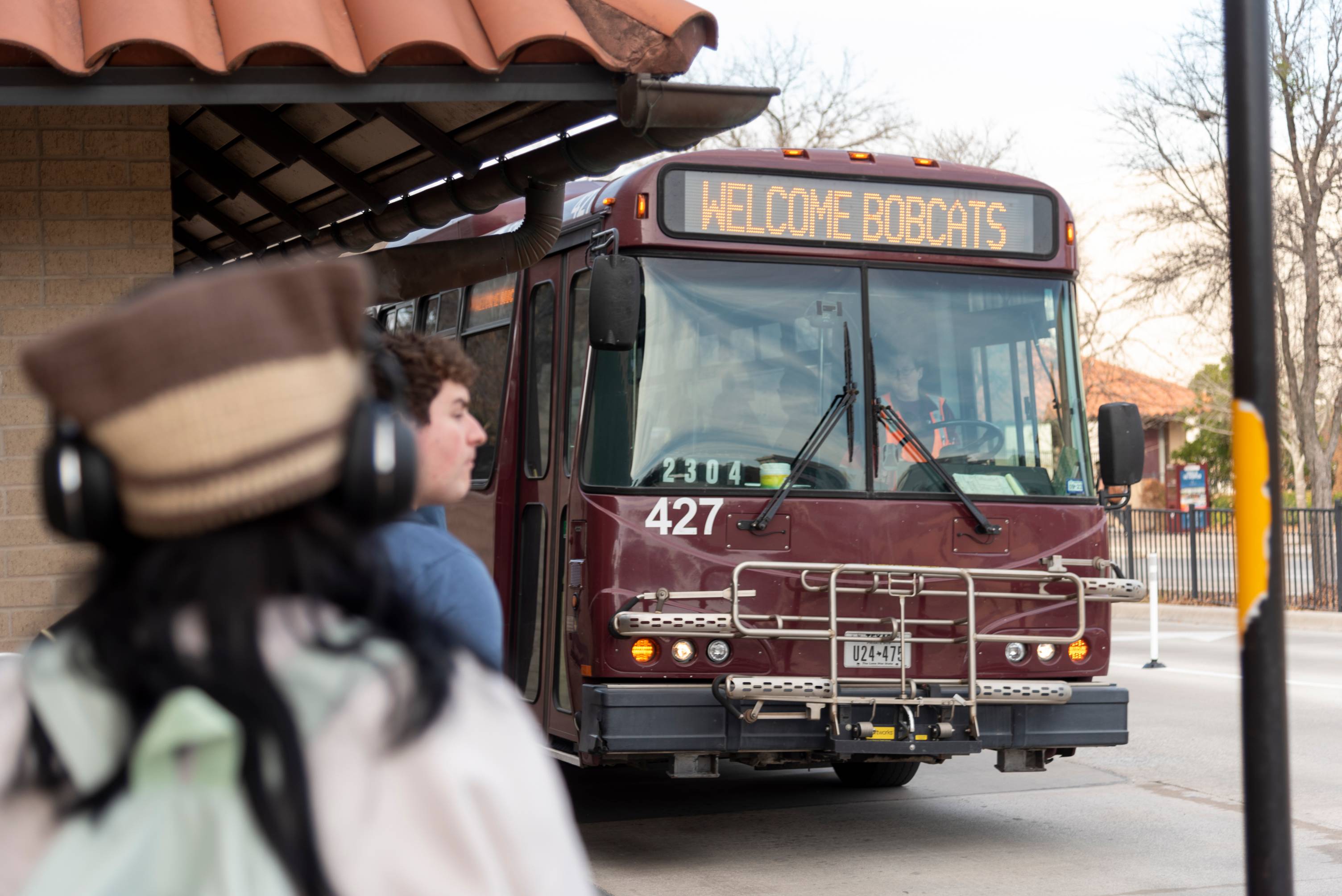 TXST shuttle approaches with a marquee reading "Welcome Bobcats". Students are awaiting the shuttles arrival.