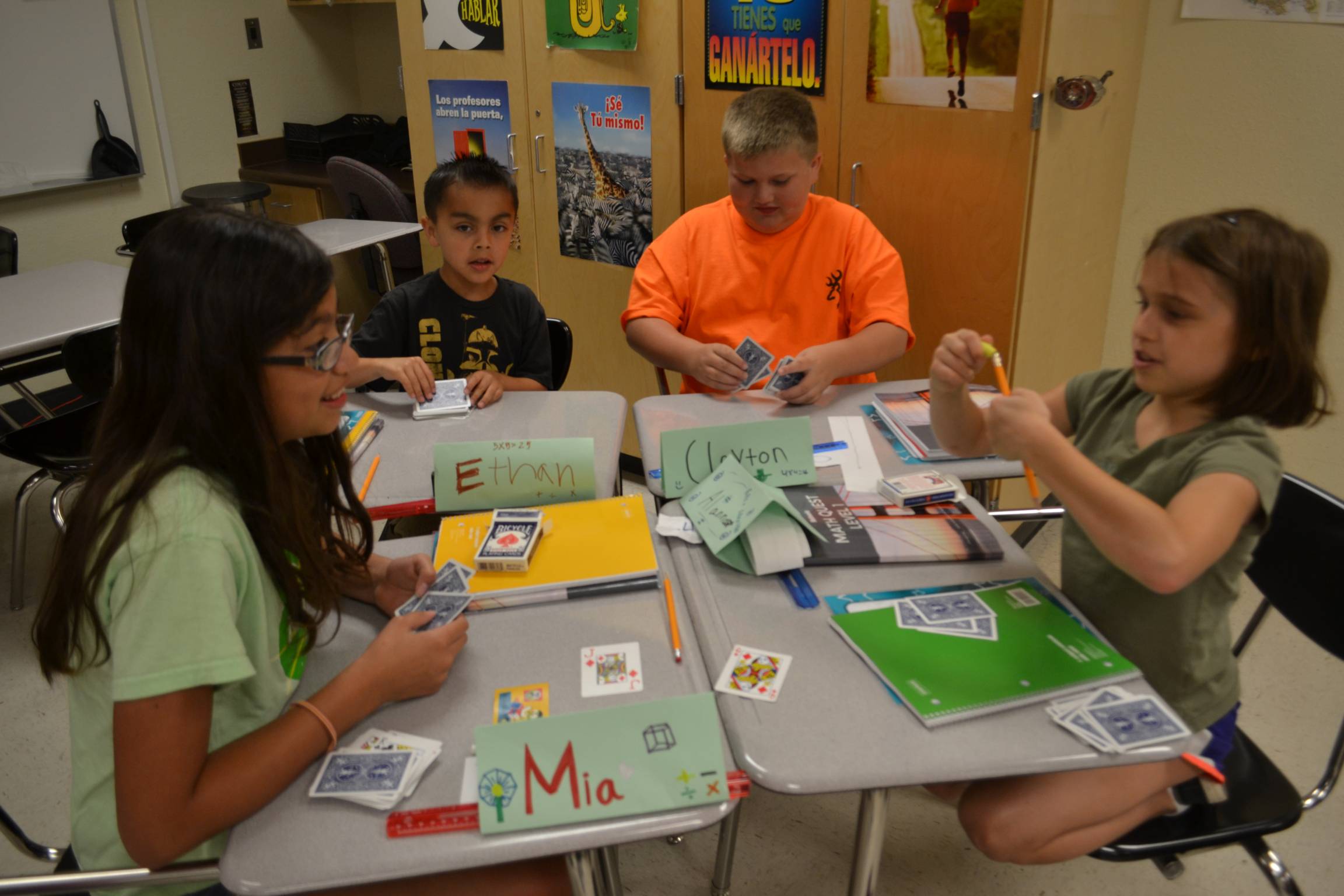 Four elementary students with desks pushed together are playing card games related to the math lesson.