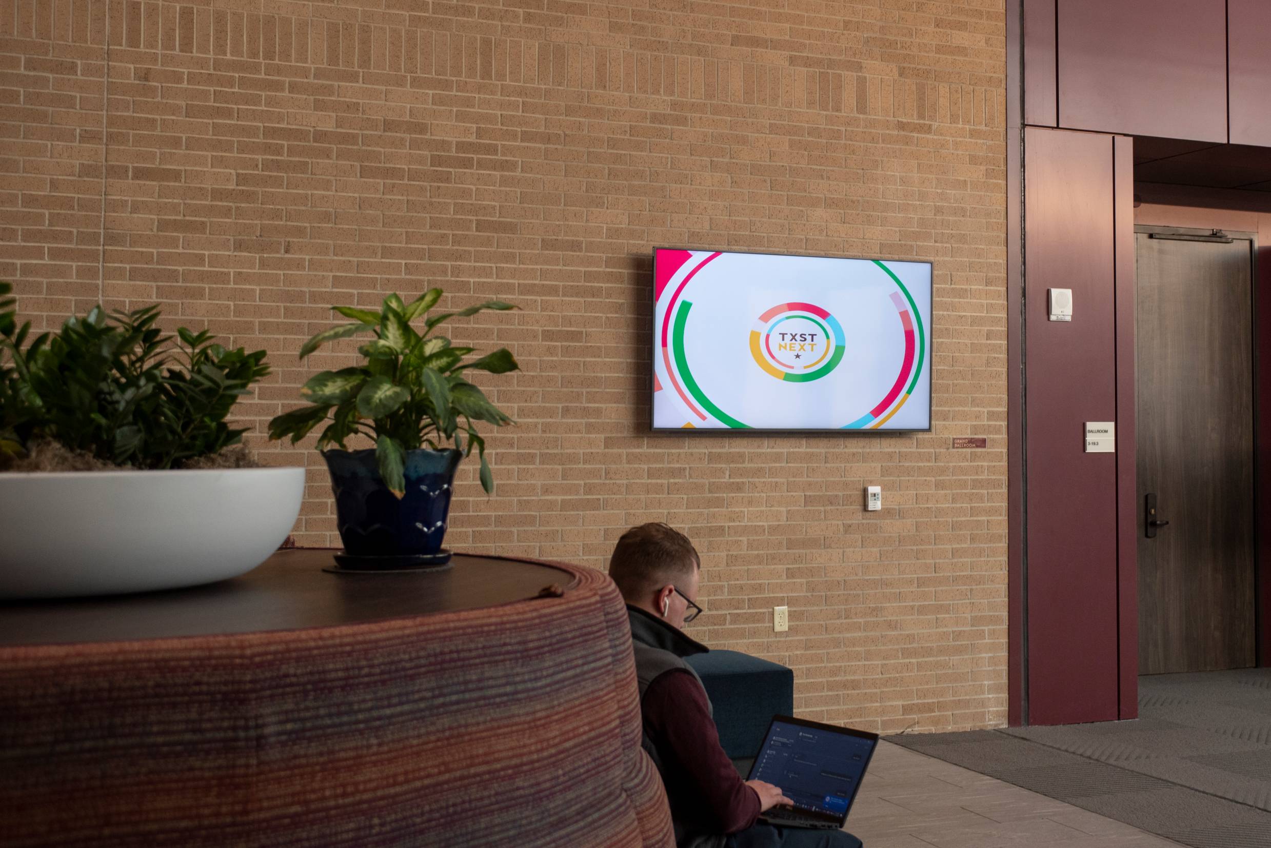 man is sitting on couch looking at a laptop with a digital board behind him hanging on a brick wall