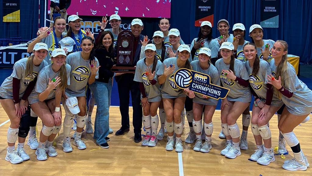 President Kelly Damphousse and First Lady Damphousse (middle) poses for a photo with the TXST volleyball team while holding the Sun Belt Conference Tournament Championship trophy.