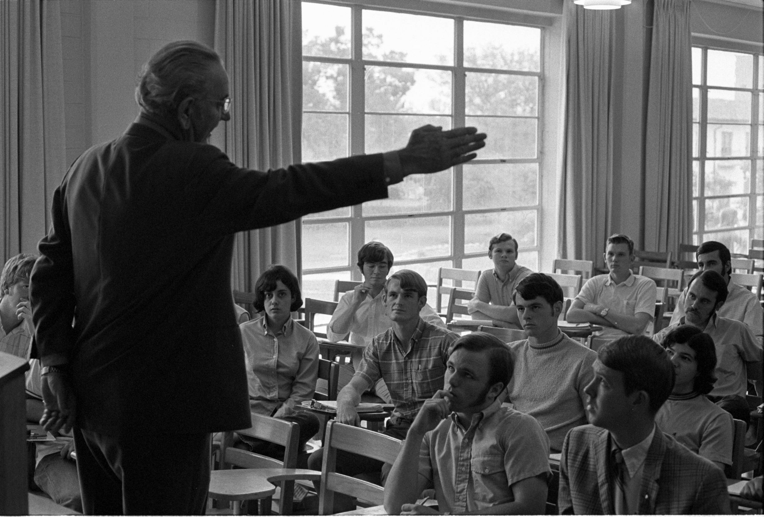 Black and white photo of LBJ in a classroom standing at the front gesturing with his right arm extended outward. The students are seated in wooden chairs appear attentive. Large windows in the background allow natural light into the room.