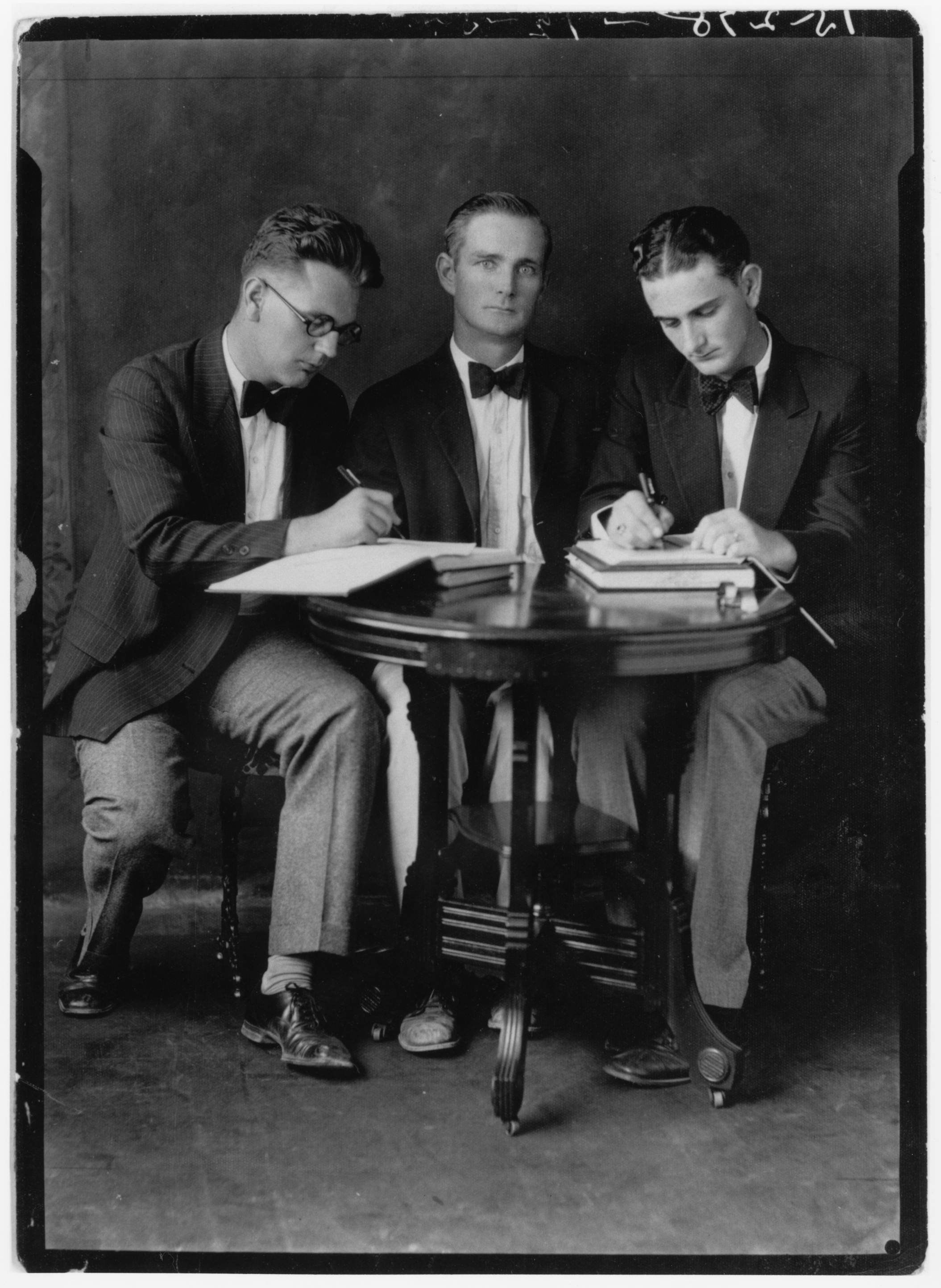 A vintage black and white photograph of LBJ seated with two other men seated around a small round table. LBJ and his classmate on the other side are focused on writing in books, while the professor in the center gazes directly at the camera. All are dressed formally in suits and bow ties, reflecting an early 20th-century style.