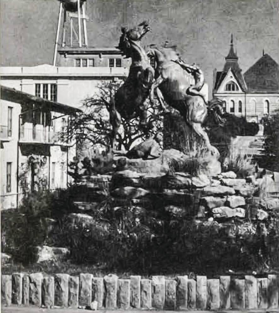 The Fighting Stallions bronze statue in its original location. On a raised dais of natural stone, surrounded by low vegetation, a pair of wild horses rear onto their hind legs. One has its head thrown back, the other is snapping in an attempt to bite. A pair of human figures are also present: one desperately clings to the back and mane of a horse, the other figure is huddled on the ground and looking back up at the fight.