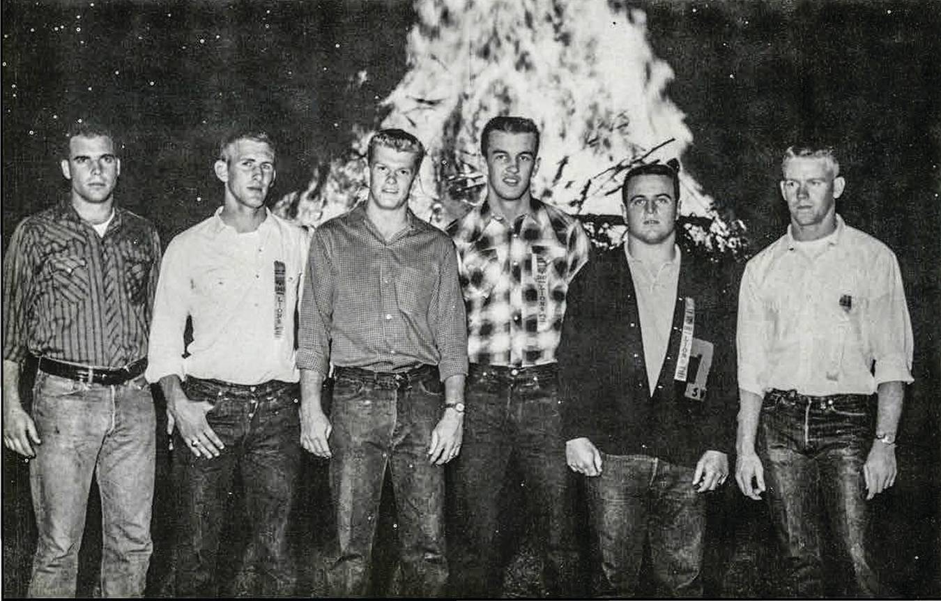 black and white archival photo from 1961. a group of 6 men stand in front of a large bonfire. 