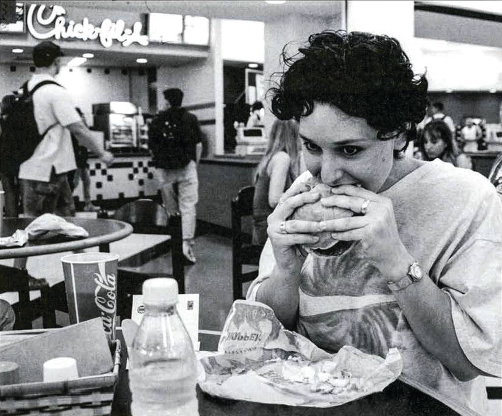 black and white photo of woman eating a sandwhich in a food court