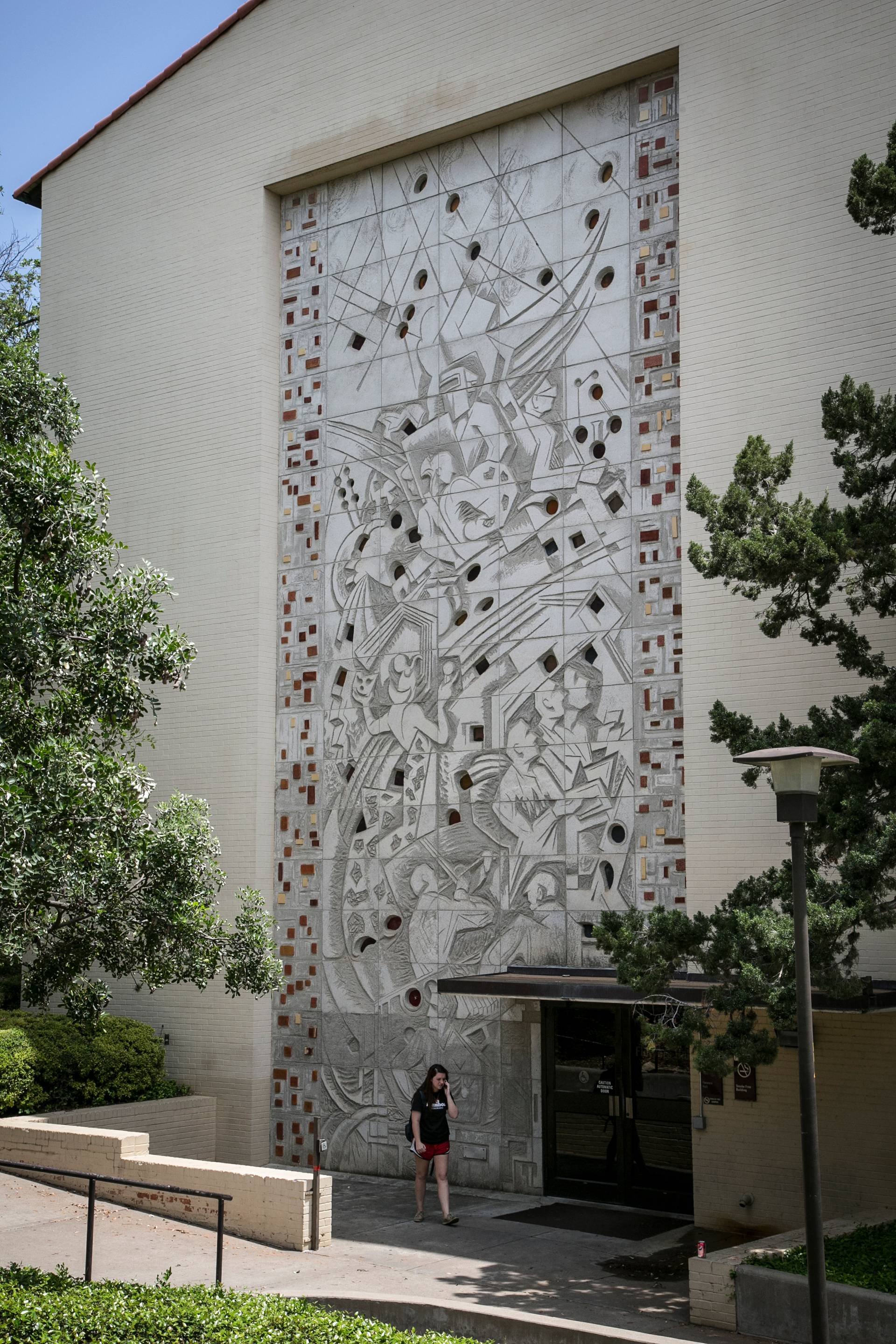 Buck Winn's Flowers Hall mural. The piece is made with concrete with small squares of colored glass inlaid throughout the composition and tiled on the left and right to make a border. The figures are carved in a geometric, abstracted style reminiscent of constructivism.